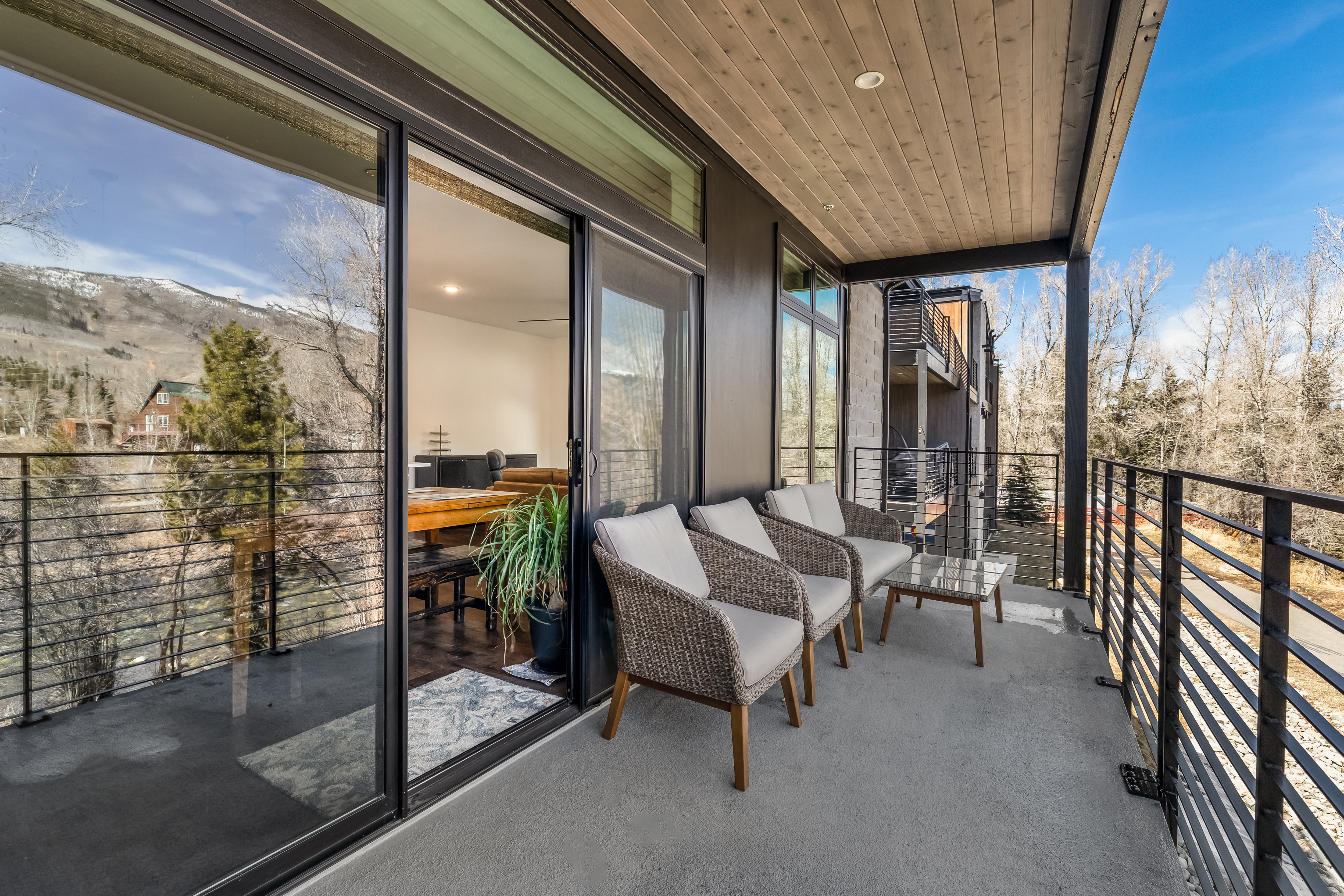 This modern balcony offers a serene outdoor living space with wicker seating and a small glass-topped table, perfect for enjoying the scenic mountain and river views. The space features a sleek metal railing, a covered wooden ceiling with recessed lighting, and large sliding glass doors that seamlessly connect the indoor dining area to the outdoors. The overall aesthetic is contemporary and inviting, emphasizing a tranquil connection to the surrounding natural landscape.