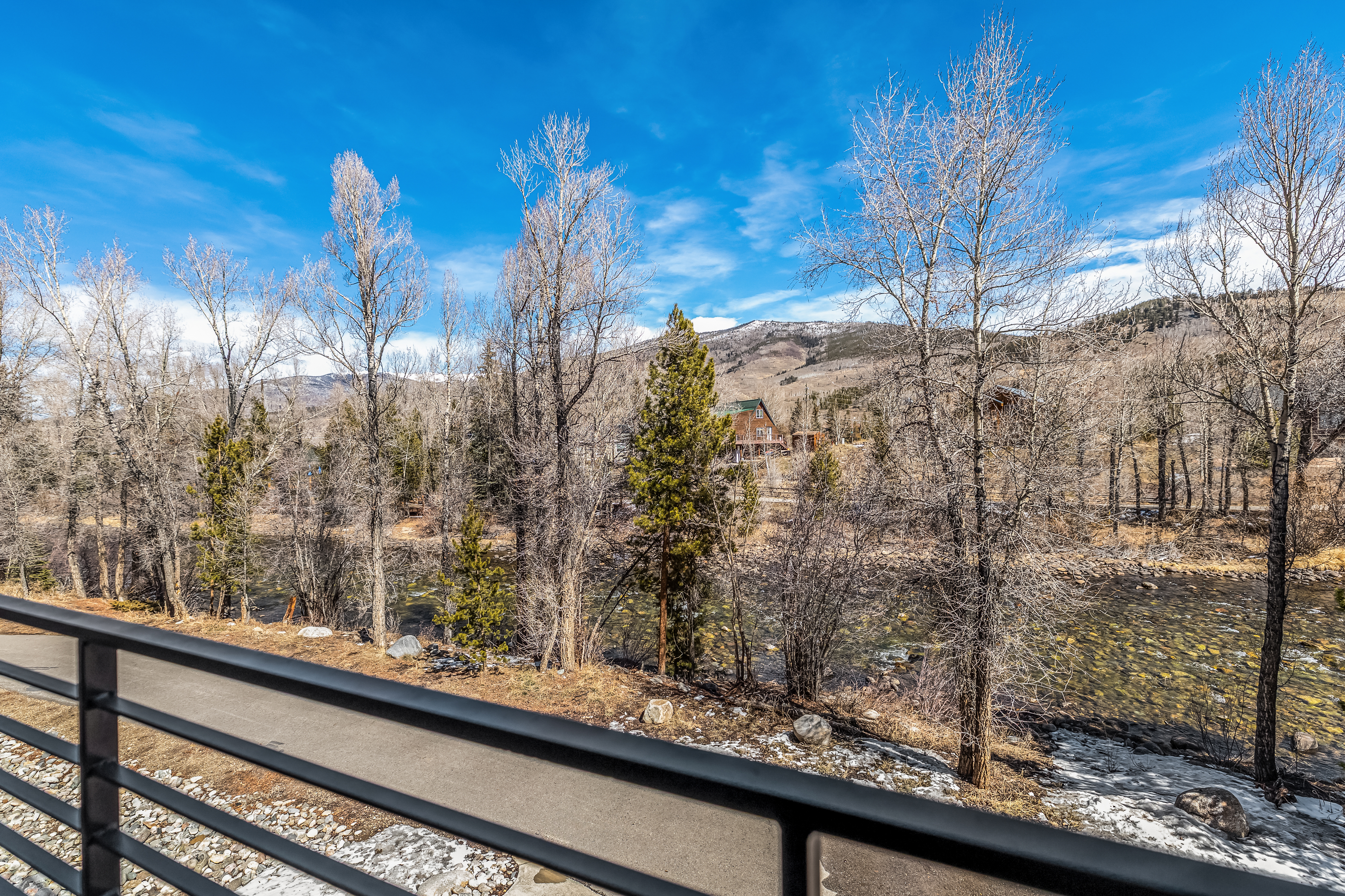 This image is captured from a balcony, offering a scenic, elevated view of a rushing river bordered by bare trees and a mountain landscape under a bright blue sky. In the distance, a rustic wooden cabin sits nestled among the trees, emphasizing the peaceful, natural setting. The foreground features a modern metal railing, providing a clear vantage point for enjoying the serene outdoor environment.