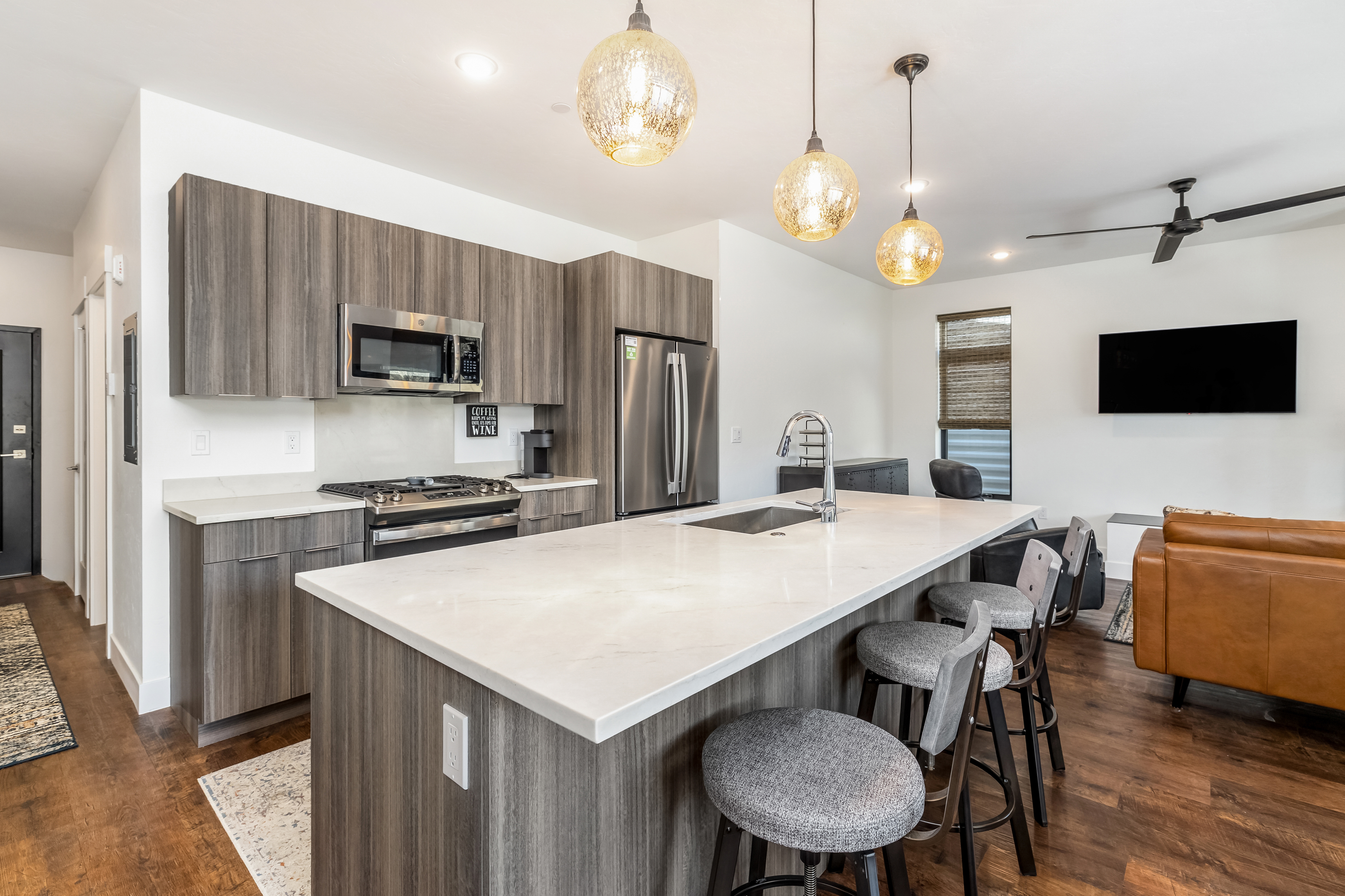 This modern, open-concept kitchen features sleek, wood-grain cabinetry paired with white quartz countertops and a spacious central island. Three textured glass pendant lights hang above the island, which includes bar seating and a stainless steel sink, while the adjacent living area creates a seamless flow for entertaining. The space is bright and contemporary, highlighted by high-end stainless steel appliances and warm wood flooring.
