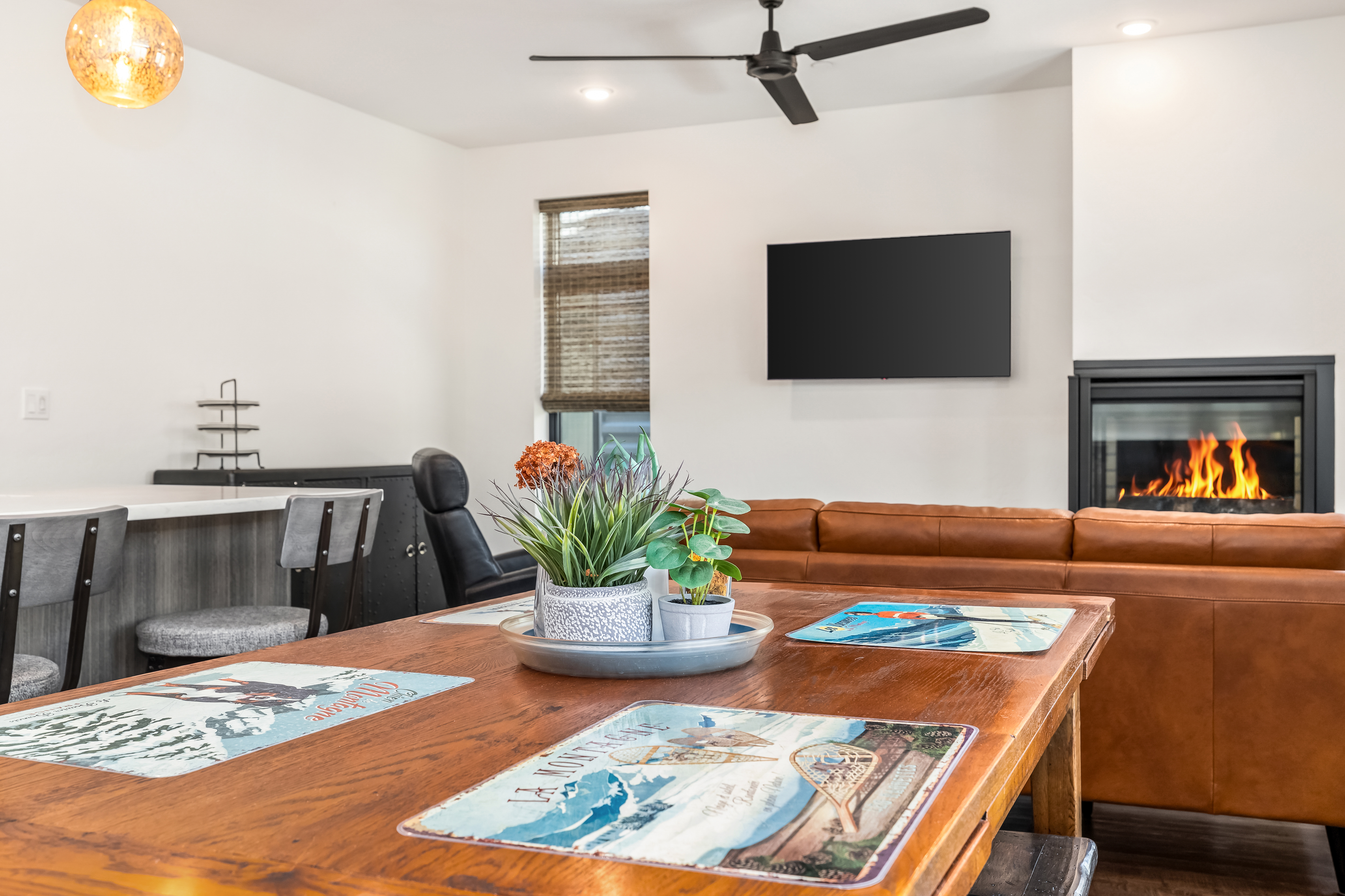 This inviting dining area features a rustic wooden table as the centerpiece, adorned with decorative placemats and a potted plant arrangement. The space seamlessly transitions into an open-concept living room, highlighted by a warm leather sofa, a cozy fireplace, and a wall-mounted television. The room is illuminated by a stylish pendant light and recessed ceiling lighting, creating a comfortable and modern atmosphere perfect for entertaining.