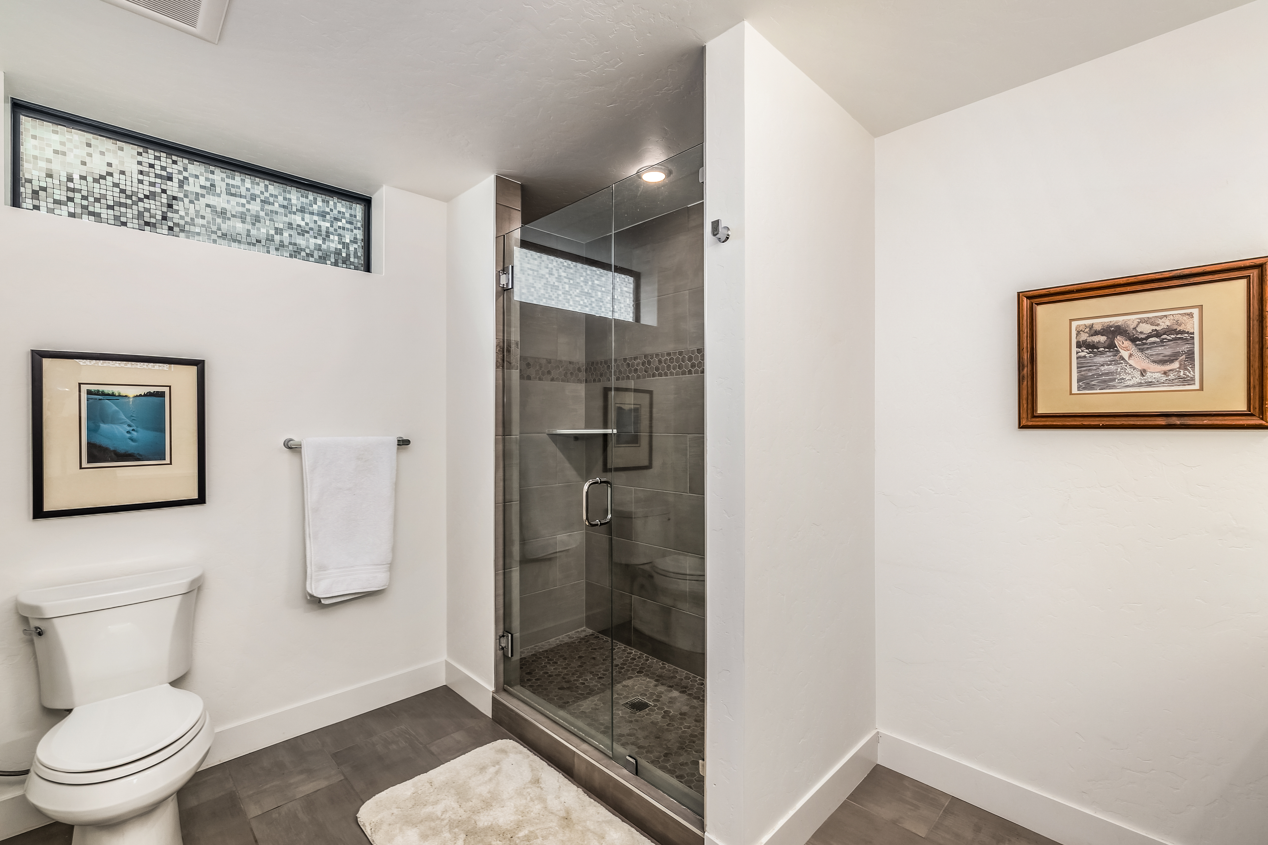 This modern bathroom features a sleek glass-enclosed walk-in shower with textured pebble flooring and neutral tile walls. The space is accented by minimalist decor, including framed artwork and a clean, white color palette that creates a bright and airy atmosphere. The perspective is a straight-on shot capturing the functional layout, including the toilet, towel rack, and shower enclosure.