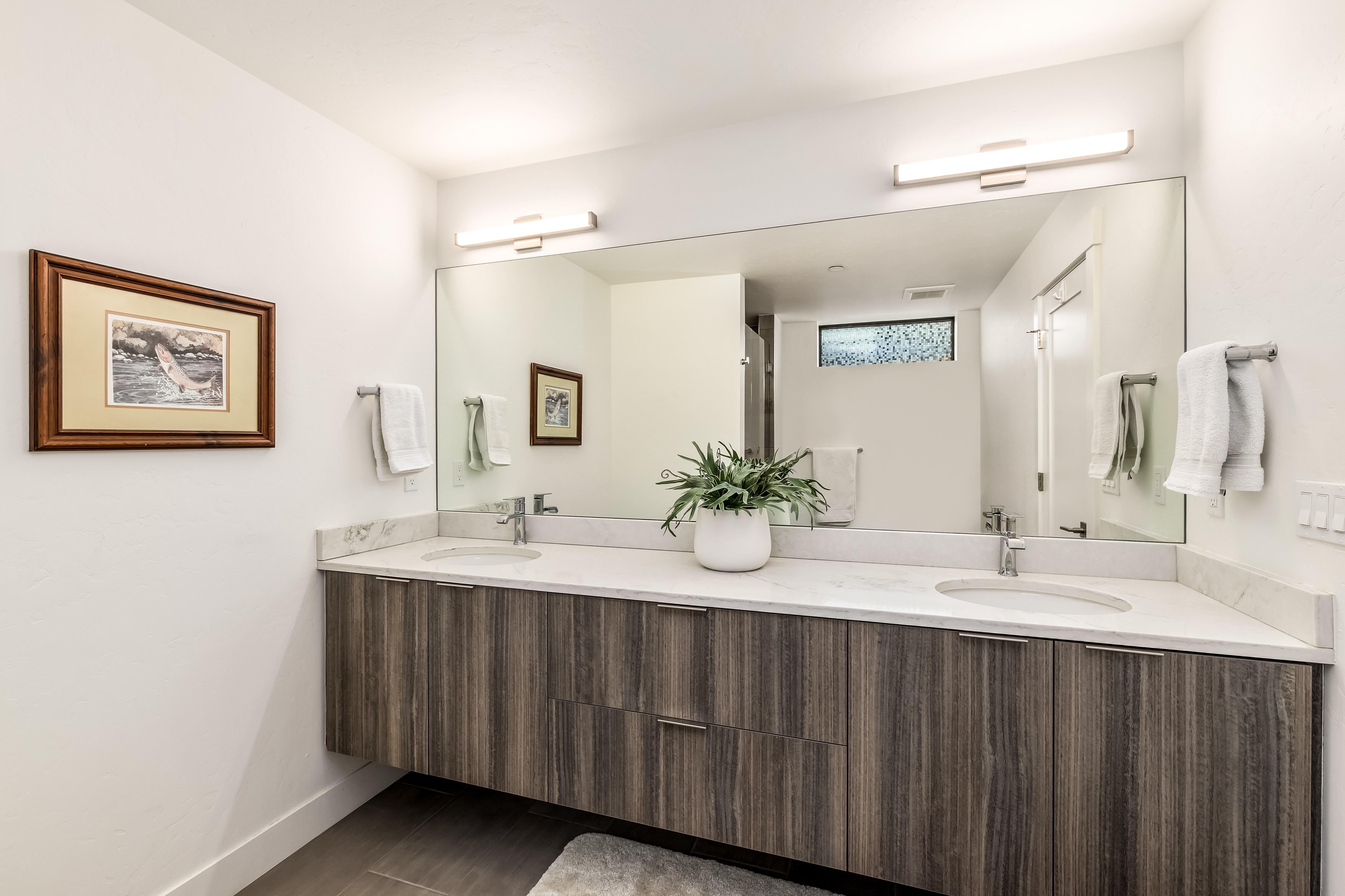 This modern primary bathroom features a sleek, floating double vanity with wood-grain cabinetry and a white marble countertop. A large, expansive mirror spans the wall, reflecting the clean lines of the space, while two minimalist light fixtures provide bright illumination. The room is accented by a framed piece of art on the left wall and a small potted plant on the vanity, creating a sophisticated and tranquil atmosphere.