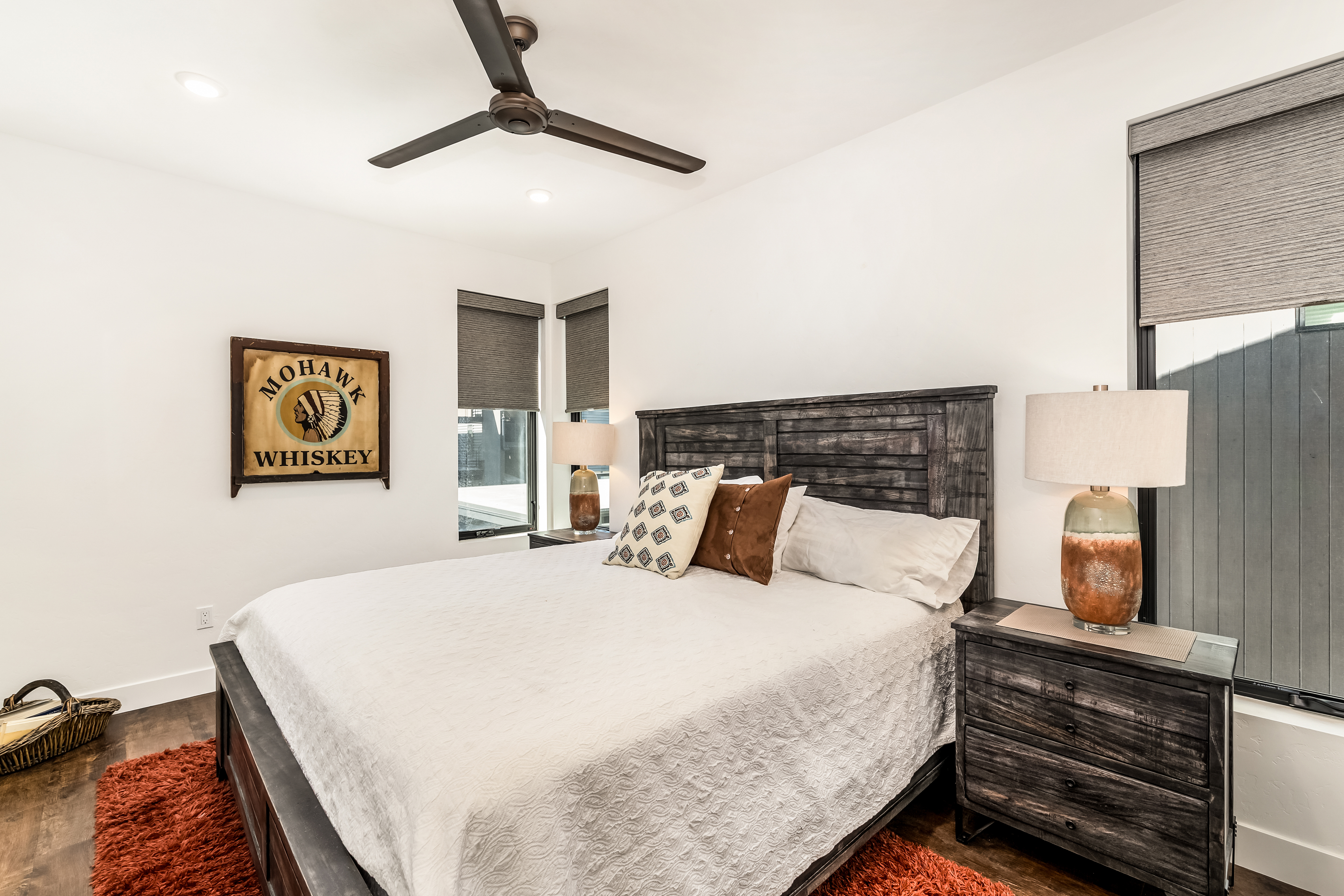 This primary bedroom features a rustic-modern aesthetic with a dark wood bed frame and matching nightstands, complemented by a textured white quilt and decorative pillows. A vibrant, shaggy orange area rug adds a pop of color to the dark hardwood floors, while a vintage-style 'Mohawk Whiskey' sign provides a unique focal point on the wall. The room is well-lit with natural light from large windows and warm bedside lamps, creating a cozy and inviting atmosphere.