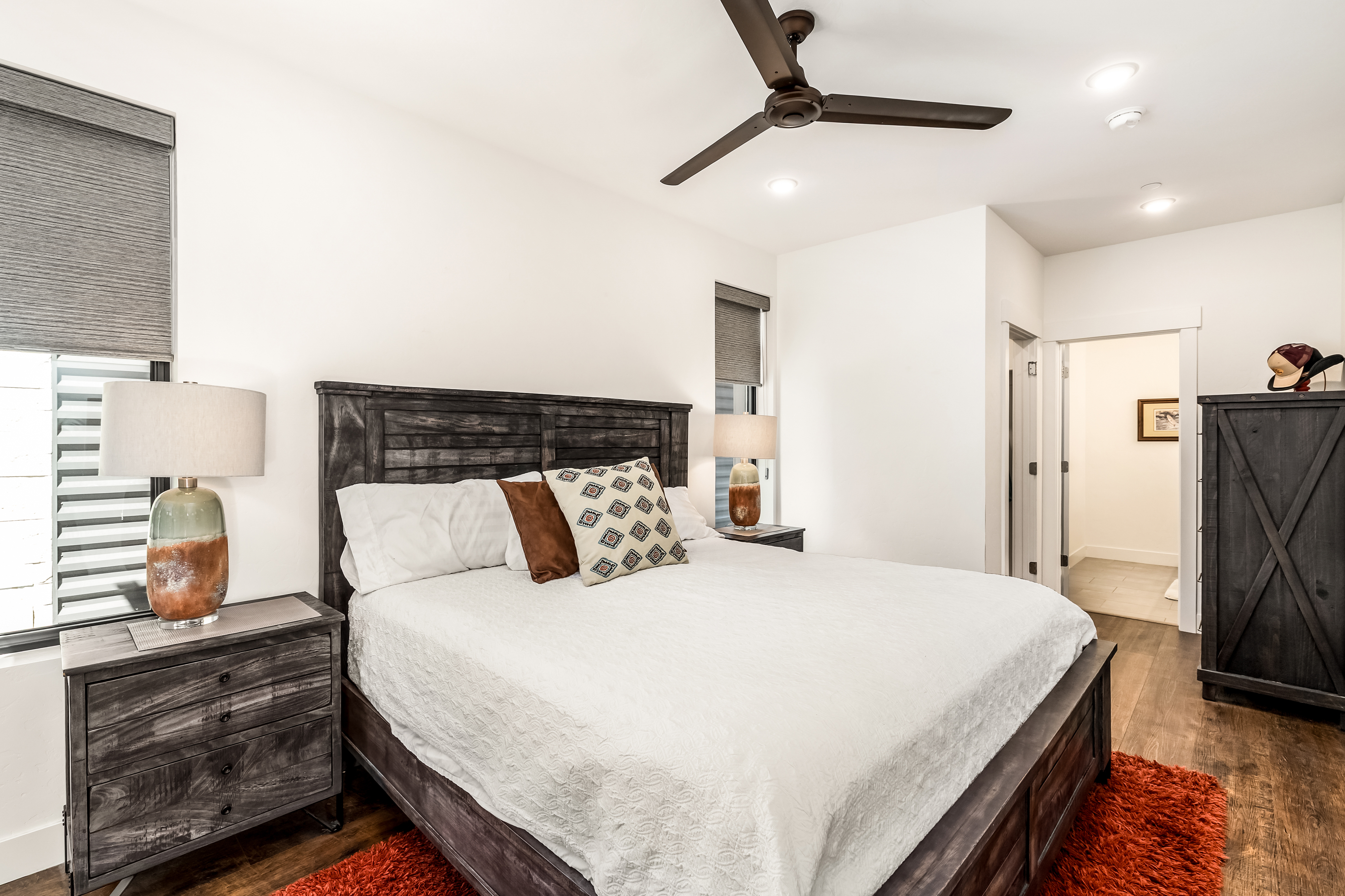 This primary bedroom features a rustic-modern aesthetic with a dark wood bed frame, matching nightstands, and a tall armoire. The room is anchored by a vibrant orange shag rug that contrasts with the neutral white bedding and walls, while a ceiling fan and recessed lighting provide a comfortable atmosphere. The perspective is a straight-on shot from the foot of the bed, highlighting the cozy, well-appointed layout and the transition into an adjacent bathroom.