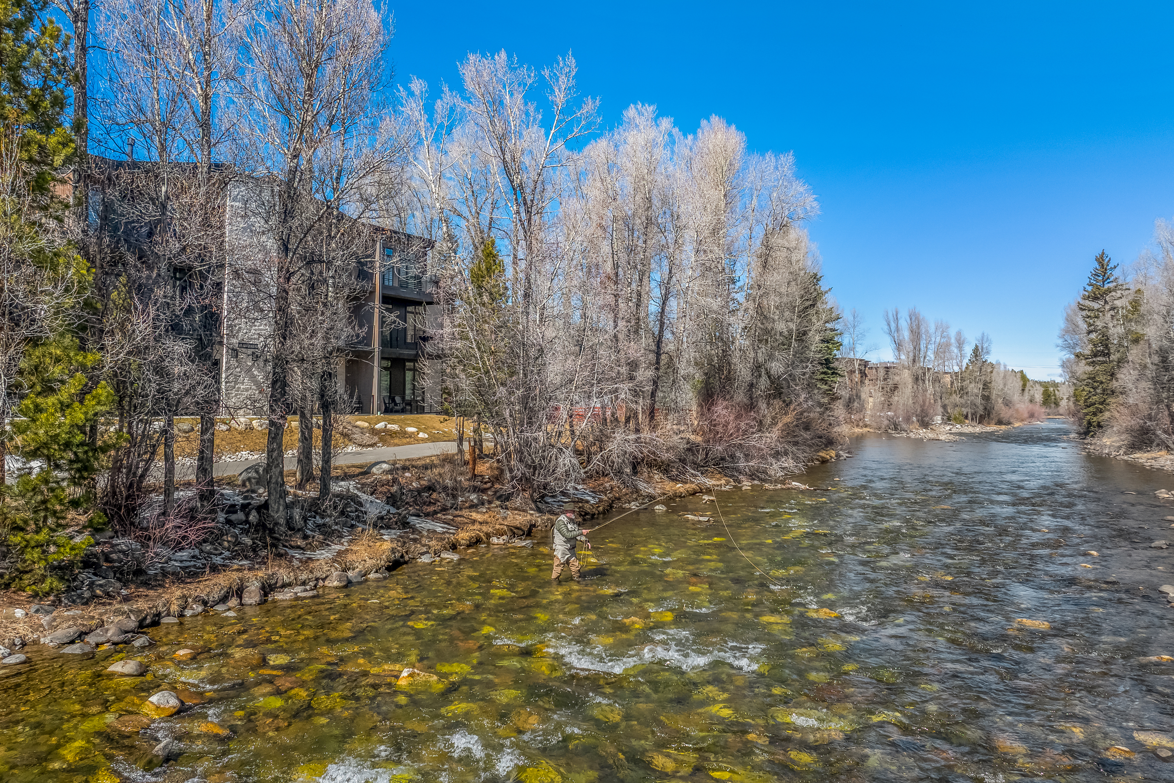 This scenic outdoor shot captures a fly fisherman wading in a clear, rocky river adjacent to a modern, multi-story residential building. The perspective is elevated, showcasing the natural beauty of the surrounding leafless trees and the tranquil, flowing water. It highlights the property's prime location, offering direct access to recreational activities and a serene, nature-focused lifestyle.