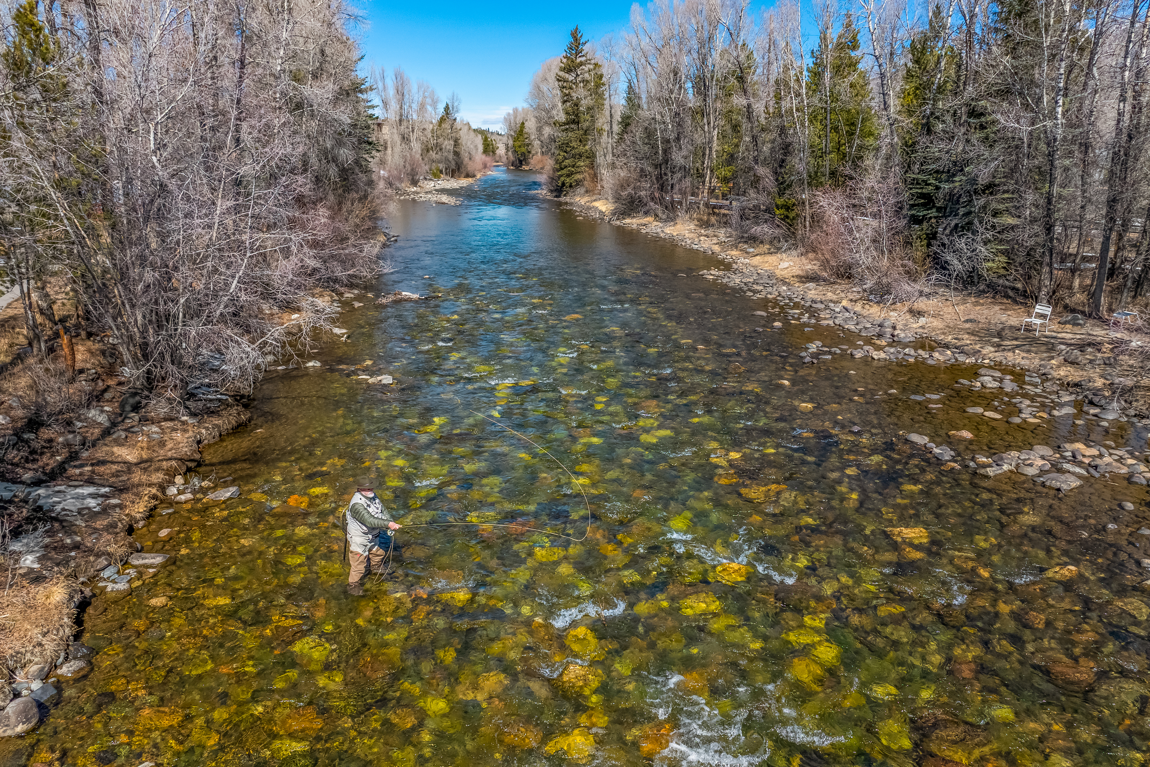 This high-angle aerial shot captures a serene, clear-water river flowing through a wooded landscape, highlighting the natural beauty and recreational potential of the property's surroundings. A fly fisherman stands in the shallow, rocky riverbed, emphasizing the peaceful and active lifestyle associated with this location. The scene is framed by tall, bare-branched trees under a bright blue sky, suggesting a tranquil and scenic outdoor environment.