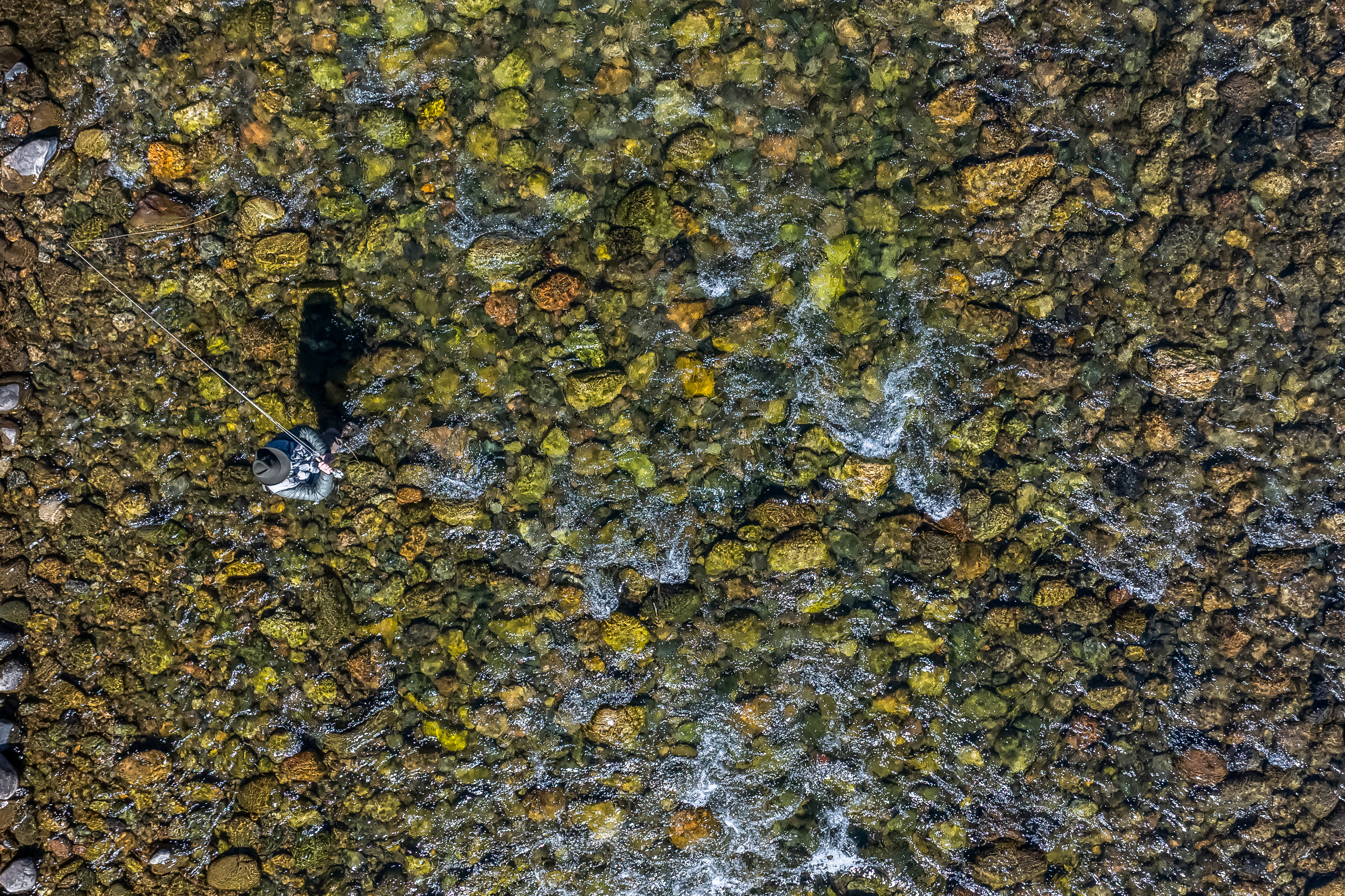 This high-angle aerial perspective captures a fly fisherman standing in a shallow, crystal-clear riverbed filled with smooth, multi-colored stones. The water flows gently around the angler, creating subtle ripples and white foam patterns that contrast with the textured river bottom. The scene evokes a sense of tranquility and outdoor recreation, highlighting the natural beauty of a pristine waterway.