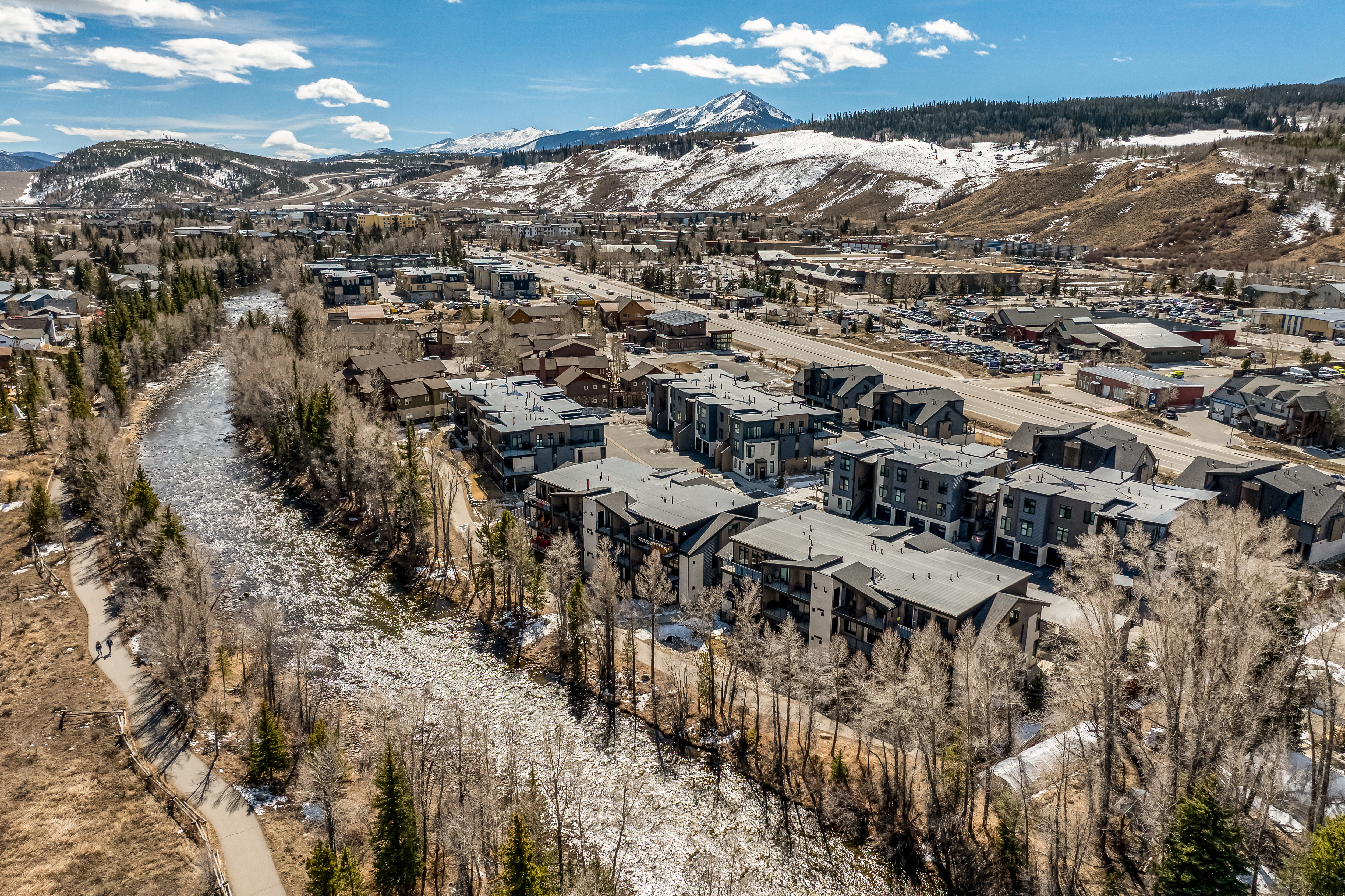 This high-angle aerial shot captures a modern residential complex nestled along a flowing river, set against a dramatic backdrop of snow-capped mountains. The scene highlights the contemporary architecture of the multi-story townhomes, a paved walking path along the water, and the surrounding mountainous landscape under a bright, clear sky. The perspective emphasizes the property's prime location, blending urban convenience with scenic natural beauty.