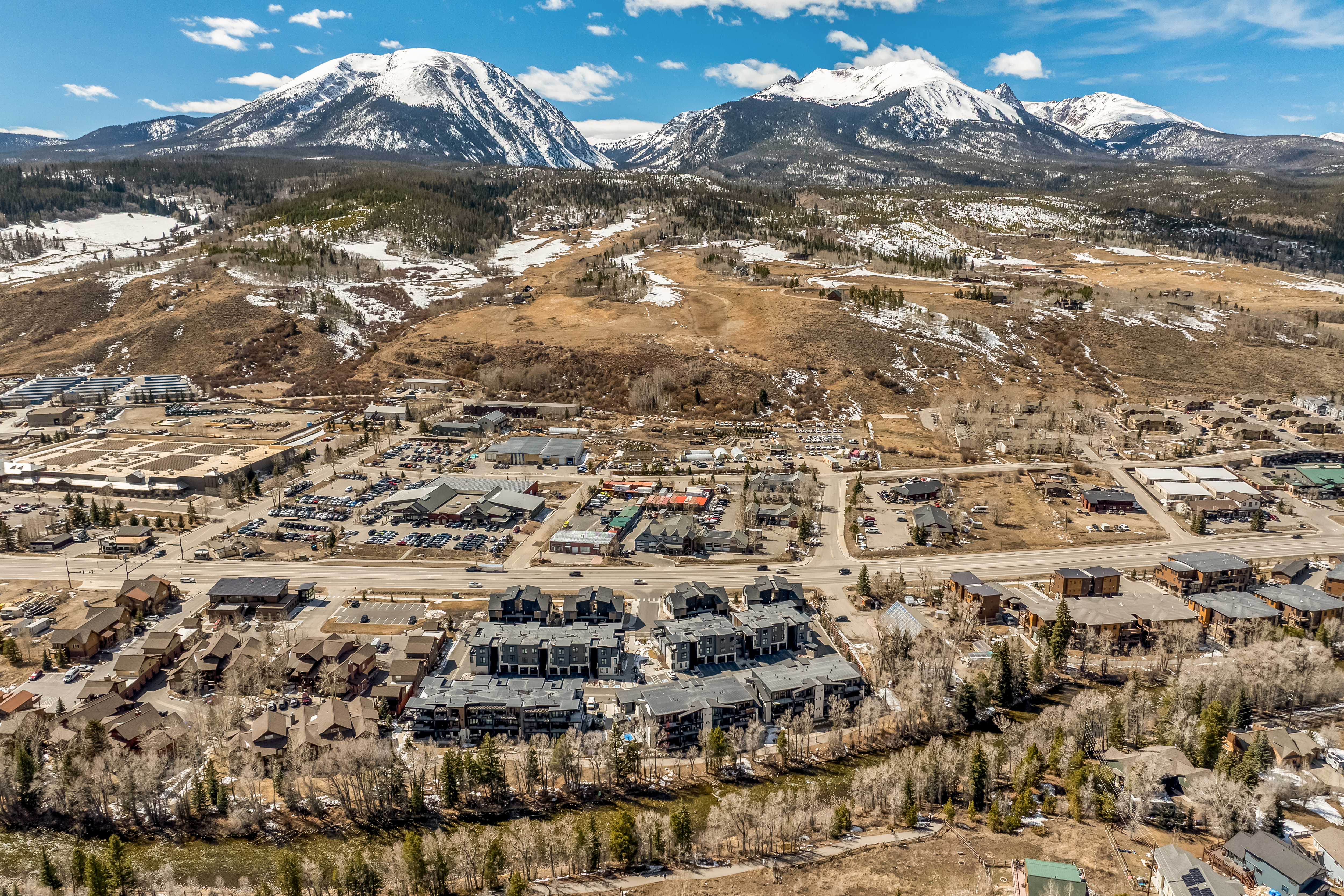 This high-altitude aerial shot captures a sprawling mountain town nestled at the base of majestic, snow-capped peaks. The foreground features modern multi-family residential complexes and commercial areas, while the mid-ground transitions into open, partially snow-dusted terrain leading up to the rugged mountain range. The perspective provides a comprehensive overview of the community's layout, highlighting its proximity to both urban amenities and the natural wilderness.