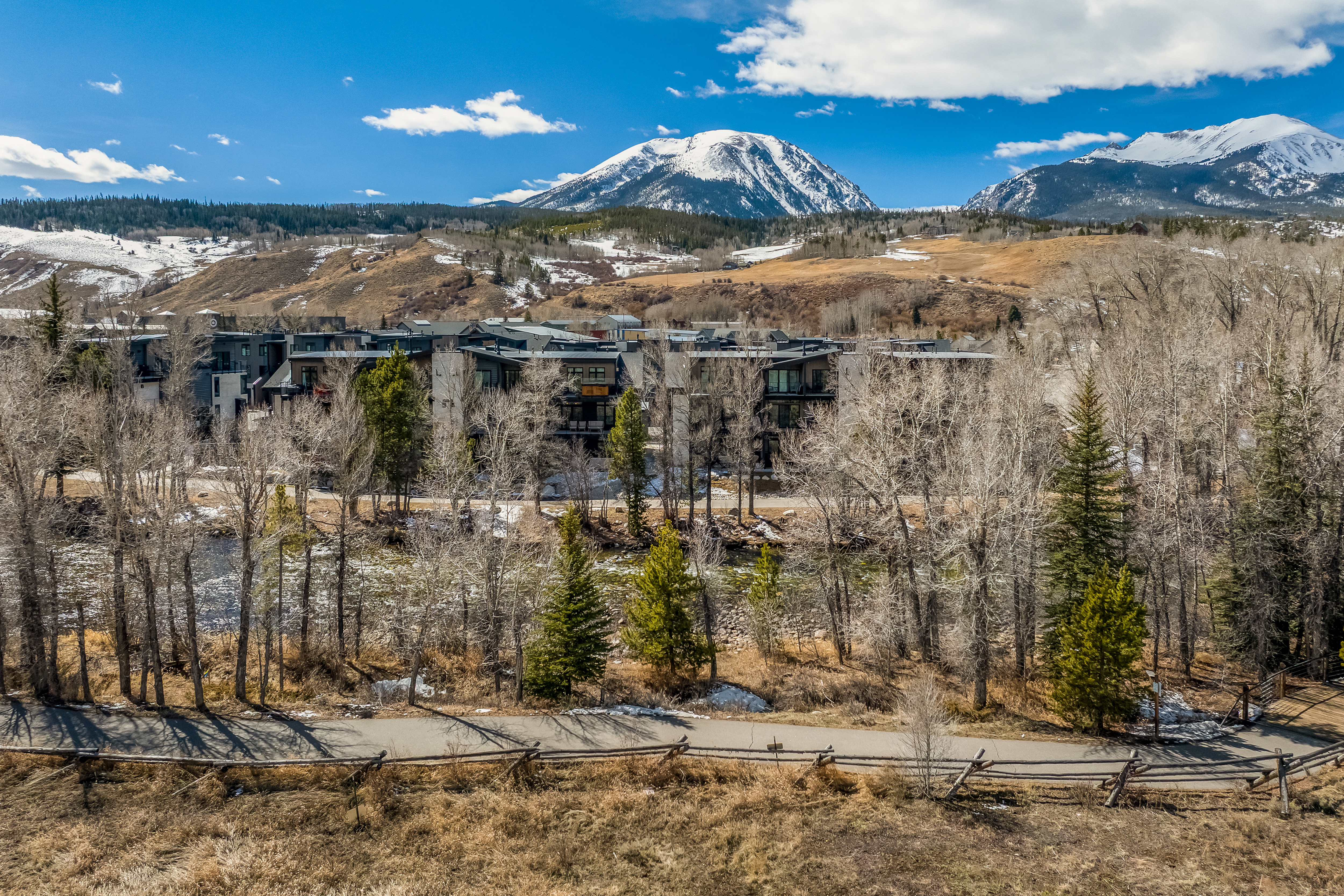 This aerial perspective captures a modern residential complex nestled in a scenic mountain landscape, featuring contemporary architecture with dark siding and large windows. In the foreground, a paved walking path winds through a natural, wooded area with a river, leading the eye toward the multi-unit buildings. The backdrop is dominated by majestic, snow-capped mountain peaks under a clear blue sky, emphasizing the property's prime location for outdoor enthusiasts.