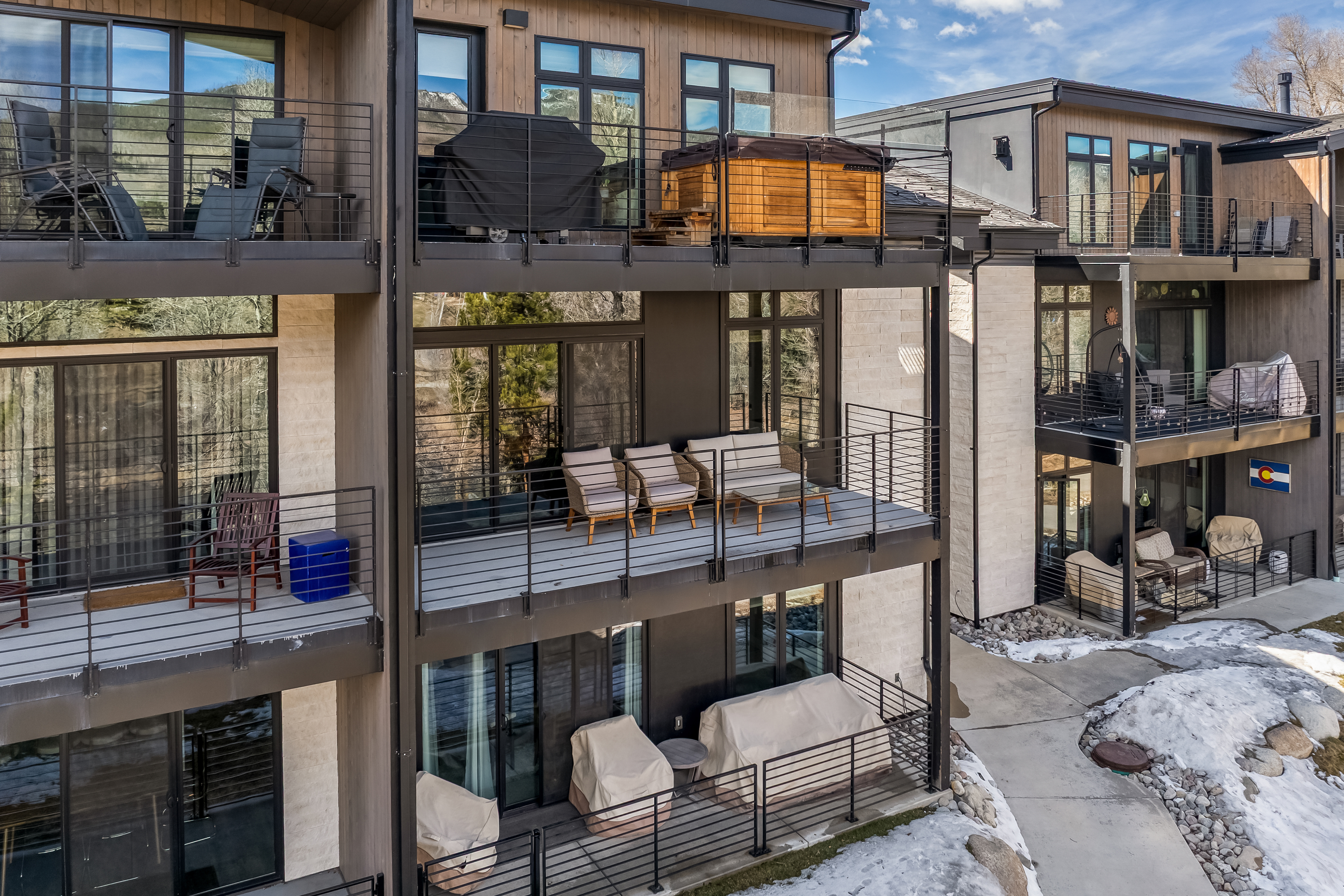 This image captures a multi-level residential building featuring stacked balconies with modern metal railings and contemporary architectural design. The balconies are furnished with outdoor seating, a hot tub, and covered furniture, showcasing a blend of luxury and functional outdoor living space. The perspective is an elevated, slightly angled exterior view that highlights the building's facade, textures, and the surrounding snowy landscape.