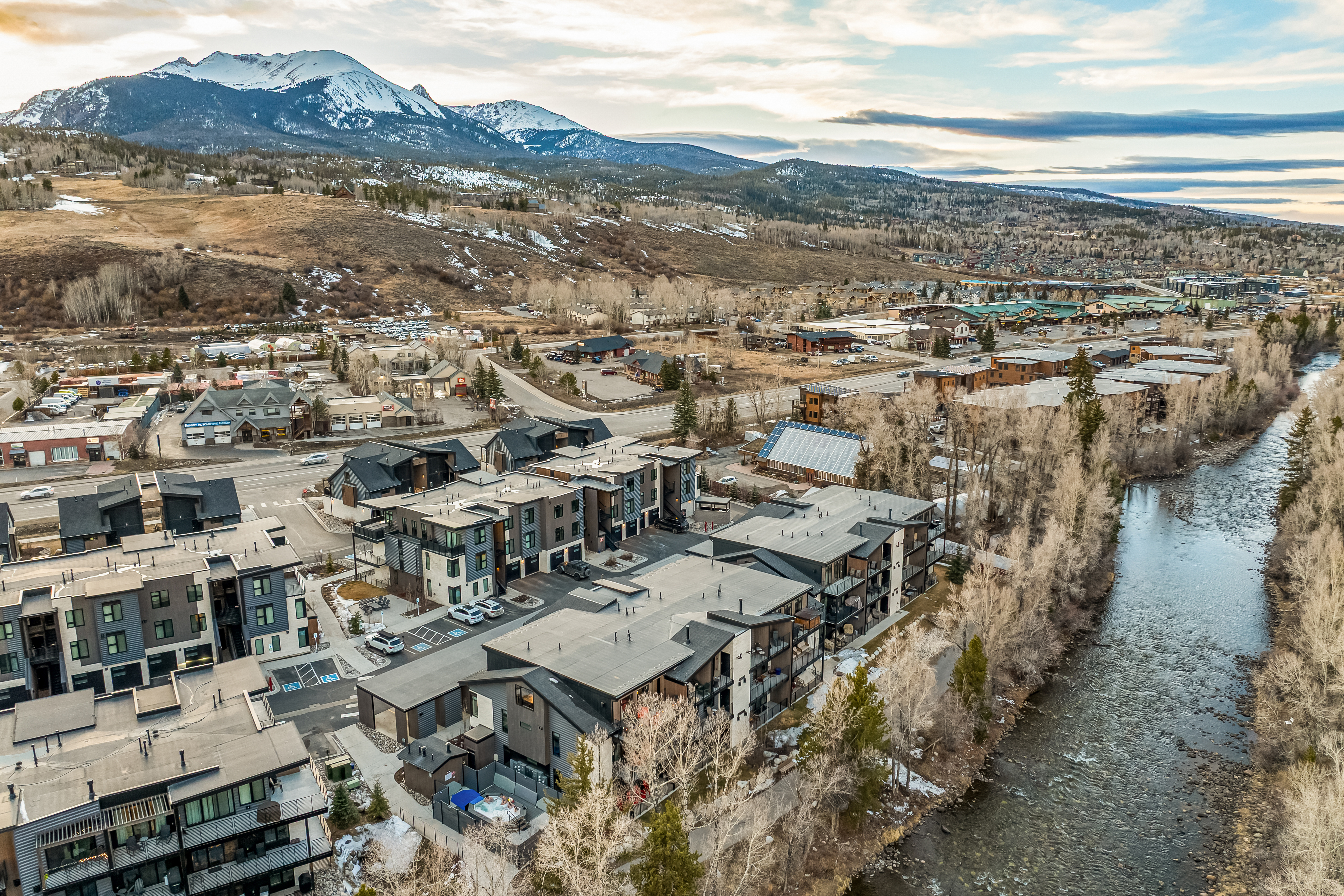 This high-angle aerial shot captures a modern residential complex nestled in a scenic mountain valley, featuring contemporary multi-story townhomes with dark metal roofing and sleek architectural lines. The property is situated directly alongside a flowing river, with a backdrop of majestic, snow-capped peaks and a clear, expansive sky. The composition highlights the integration of modern living spaces with the natural beauty of the surrounding landscape.