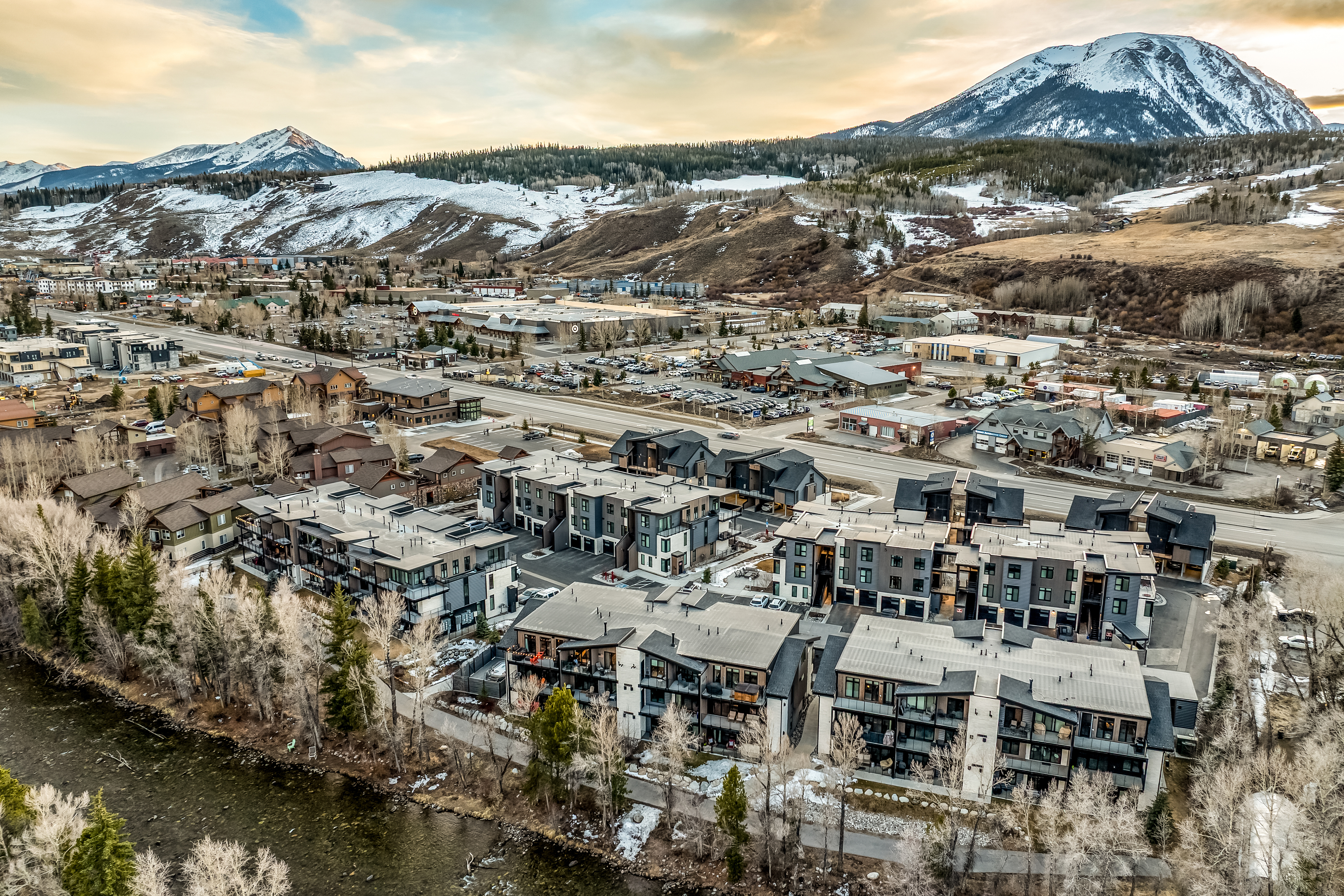 This high-angle aerial shot captures a modern residential complex nestled along a riverbank, set against a breathtaking backdrop of snow-capped mountains. The architecture features contemporary multi-story townhomes with dark siding and balconies, integrated into a scenic landscape with sparse trees and nearby commercial developments. The perspective provides a comprehensive view of the property's location, highlighting its proximity to both nature and local amenities.