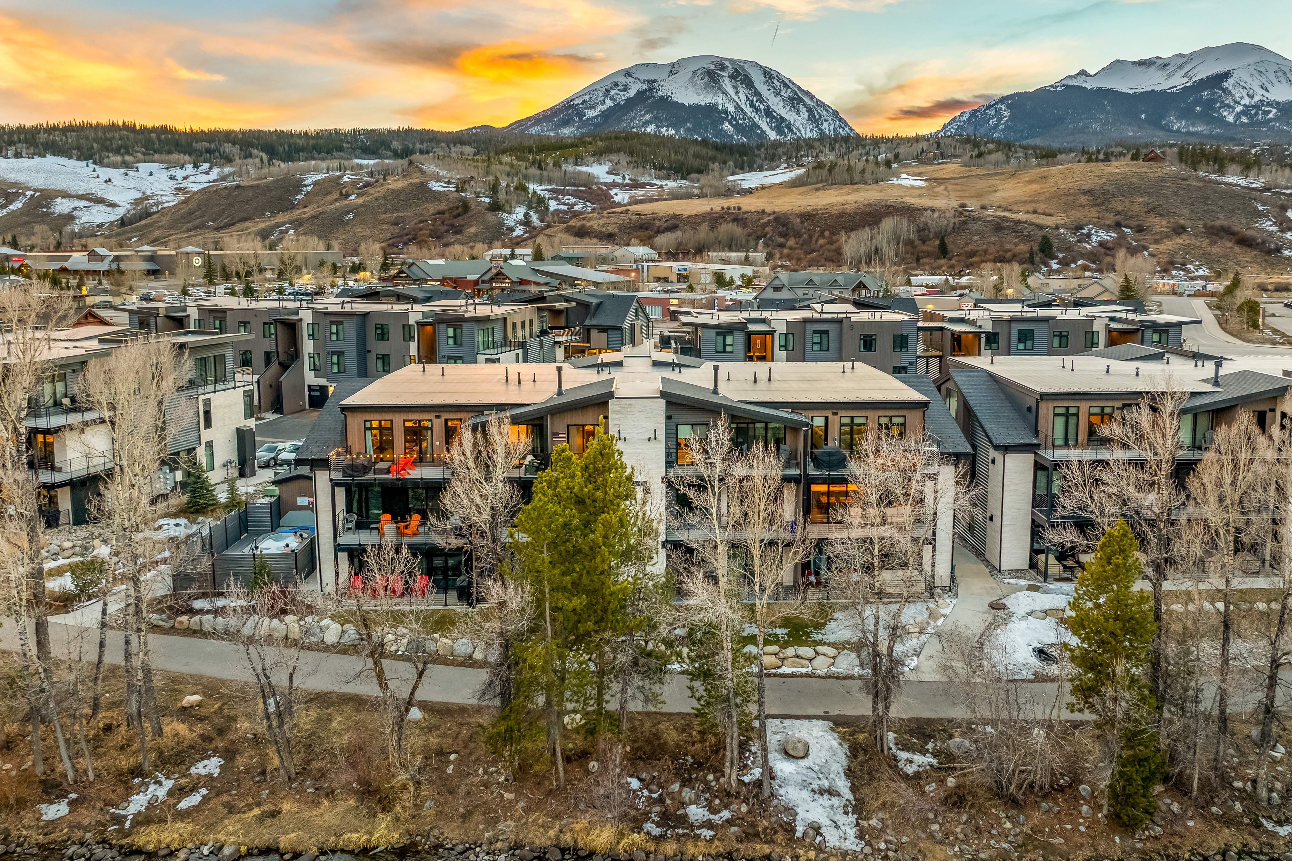 This stunning aerial view captures a modern multi-unit residential complex nestled in a mountainous landscape during a vibrant sunset. The architecture features clean lines, contemporary materials, and private balconies, with a scenic walking path and a small river running along the foreground. The dramatic backdrop of snow-capped peaks and a golden, cloud-filled sky creates an inviting and upscale atmosphere.