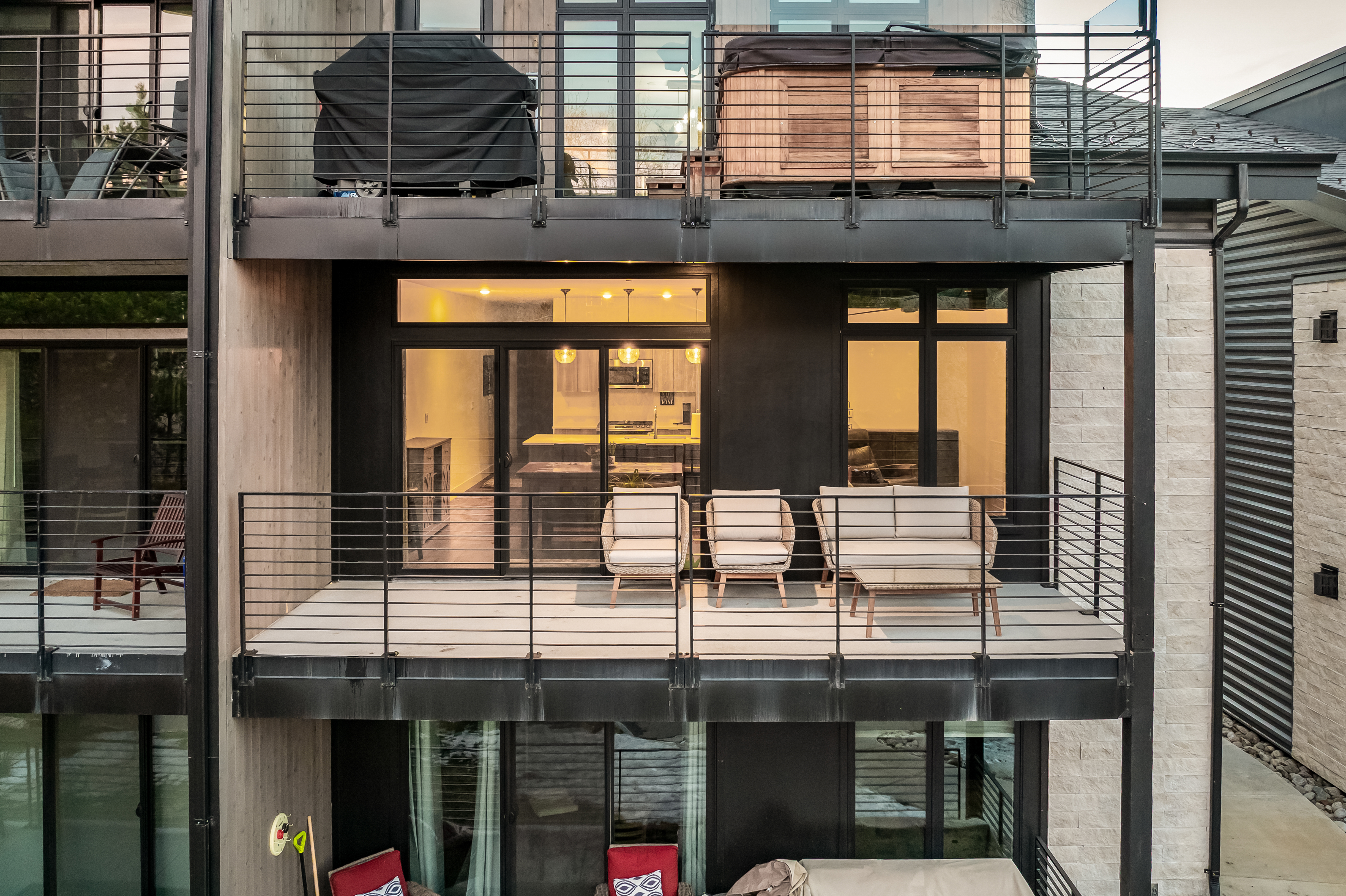This image captures a multi-level balcony view of a modern residential building, showcasing a spacious deck furnished with contemporary outdoor seating. The scene highlights a seamless transition between the indoor living area and the exterior space, featuring sleek black metal railings and warm interior lighting that creates an inviting atmosphere. The architectural design emphasizes clean lines and a sophisticated, urban aesthetic.