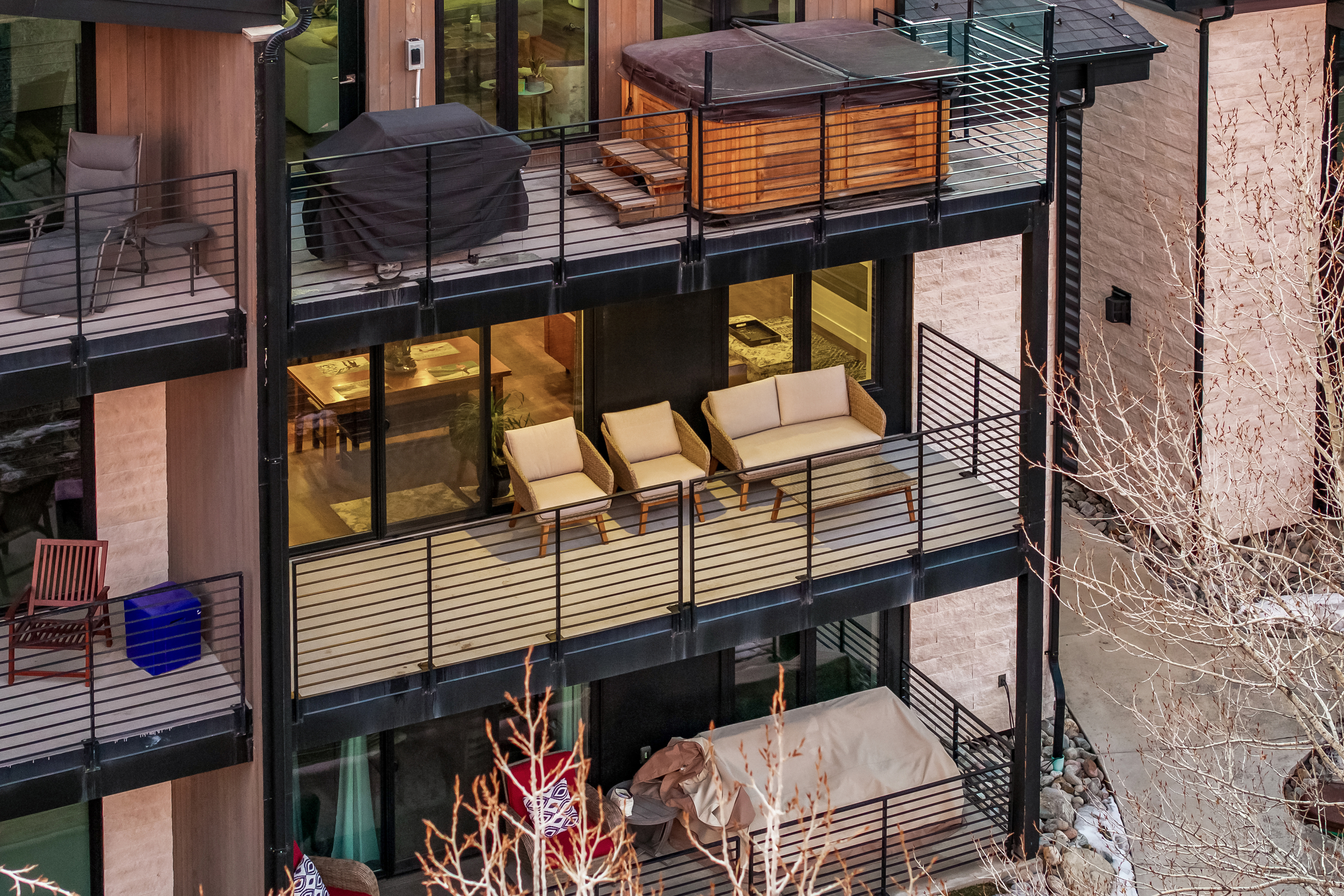This high-angle, cinematic shot captures a multi-level residential building featuring stacked balconies with modern black metal railings. The top balcony showcases a luxurious hot tub and a covered grill, while the middle level is furnished with comfortable outdoor seating, and the lower level includes a covered area. The architectural style is contemporary and sleek, emphasizing outdoor living spaces that overlook a natural, wooded setting.