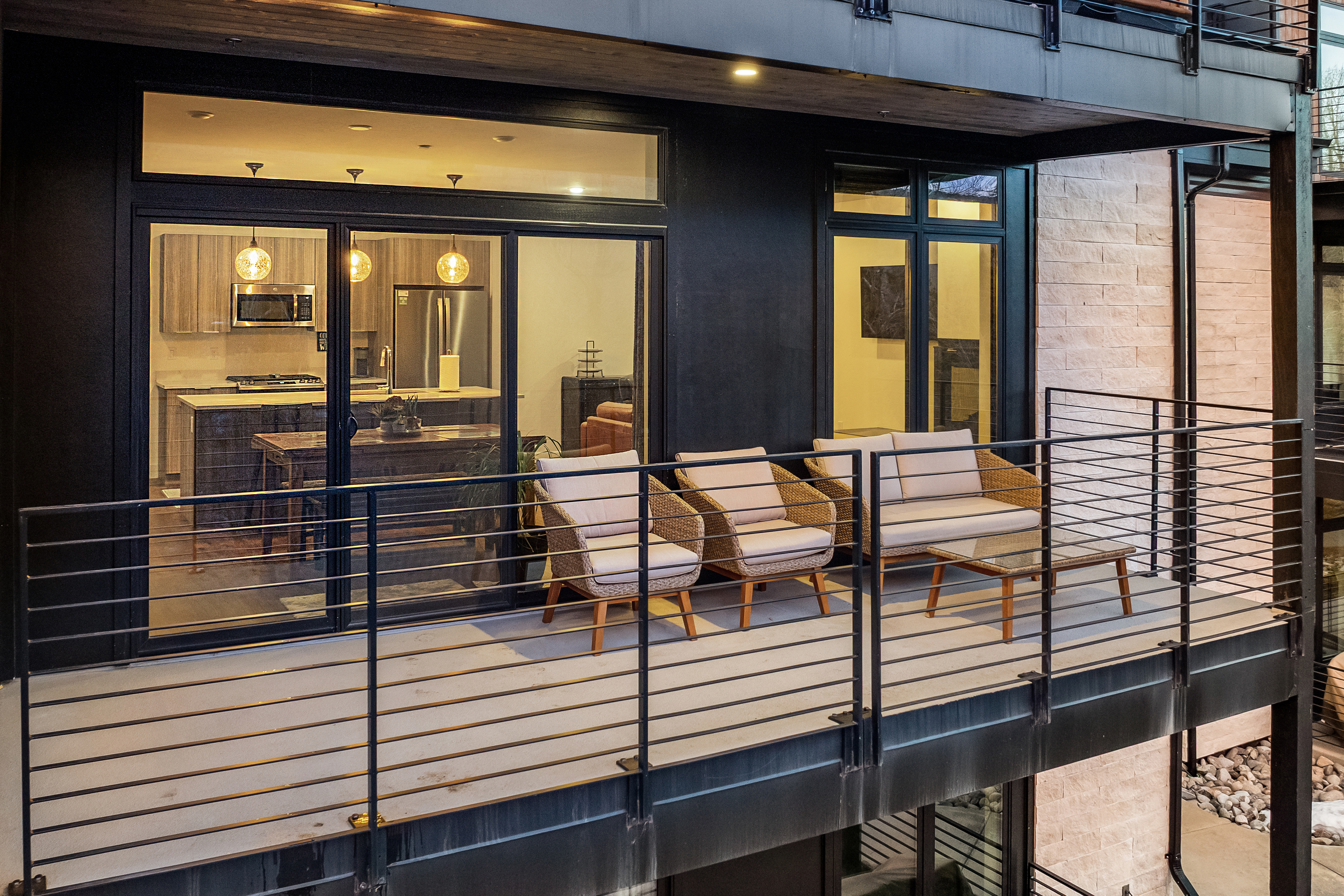 This modern balcony features a sleek, industrial-style metal railing and a spacious concrete deck furnished with comfortable wicker seating and a small coffee table. Through the large glass sliding doors, a well-appointed kitchen with warm pendant lighting and stainless steel appliances is visible, creating a seamless transition between indoor and outdoor living. The overall aesthetic is contemporary and sophisticated, perfect for relaxing or entertaining in an urban setting.