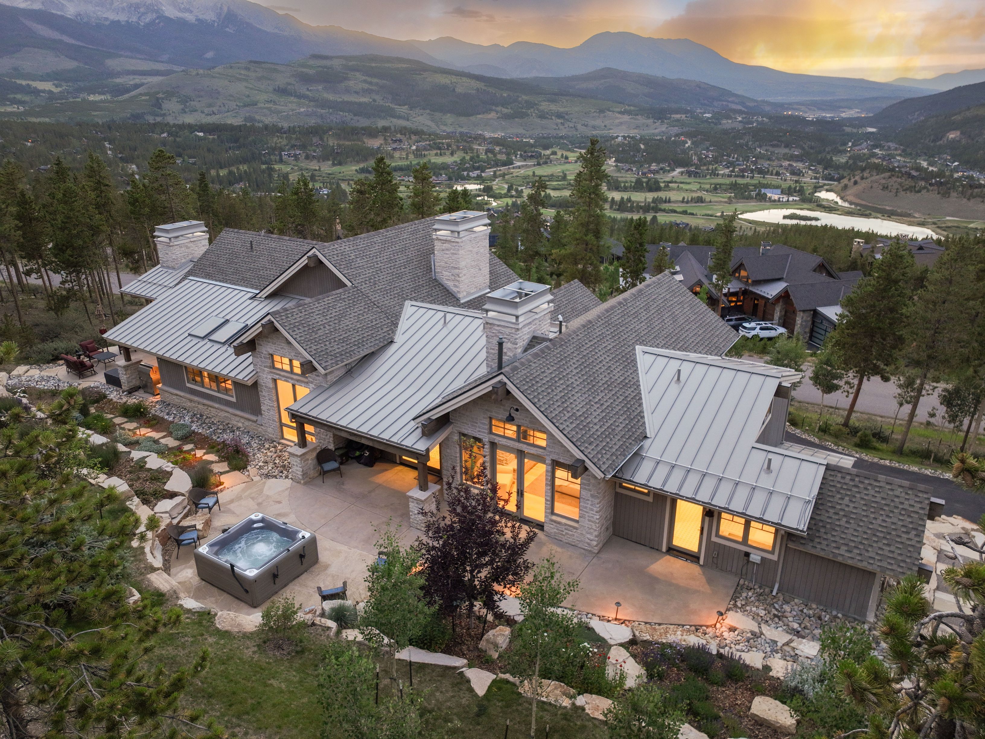 This high-angle aerial view captures a luxurious mountain residence nestled in a scenic, elevated landscape. The home features a sophisticated blend of stone and metal roofing, with a spacious concrete patio area that includes a built-in hot tub and outdoor seating. The property is surrounded by evergreen trees and offers expansive, breathtaking views of the valley, lake, and distant mountain ranges at sunset.