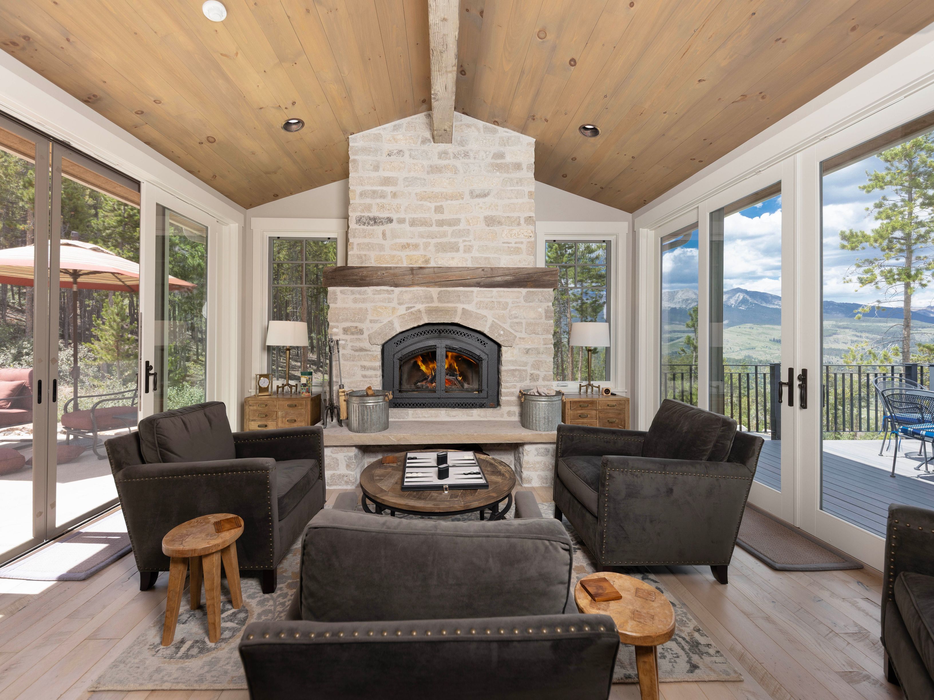 This inviting living room features a prominent stone-clad chimney breast with a central fireplace and a rustic wood mantel. The space is framed by large glass doors on either side that provide expansive views of the surrounding forest and mountain landscape. With its vaulted wood-paneled ceiling, comfortable dark upholstered seating, and a central round coffee table, the room offers a cozy and sophisticated atmosphere.