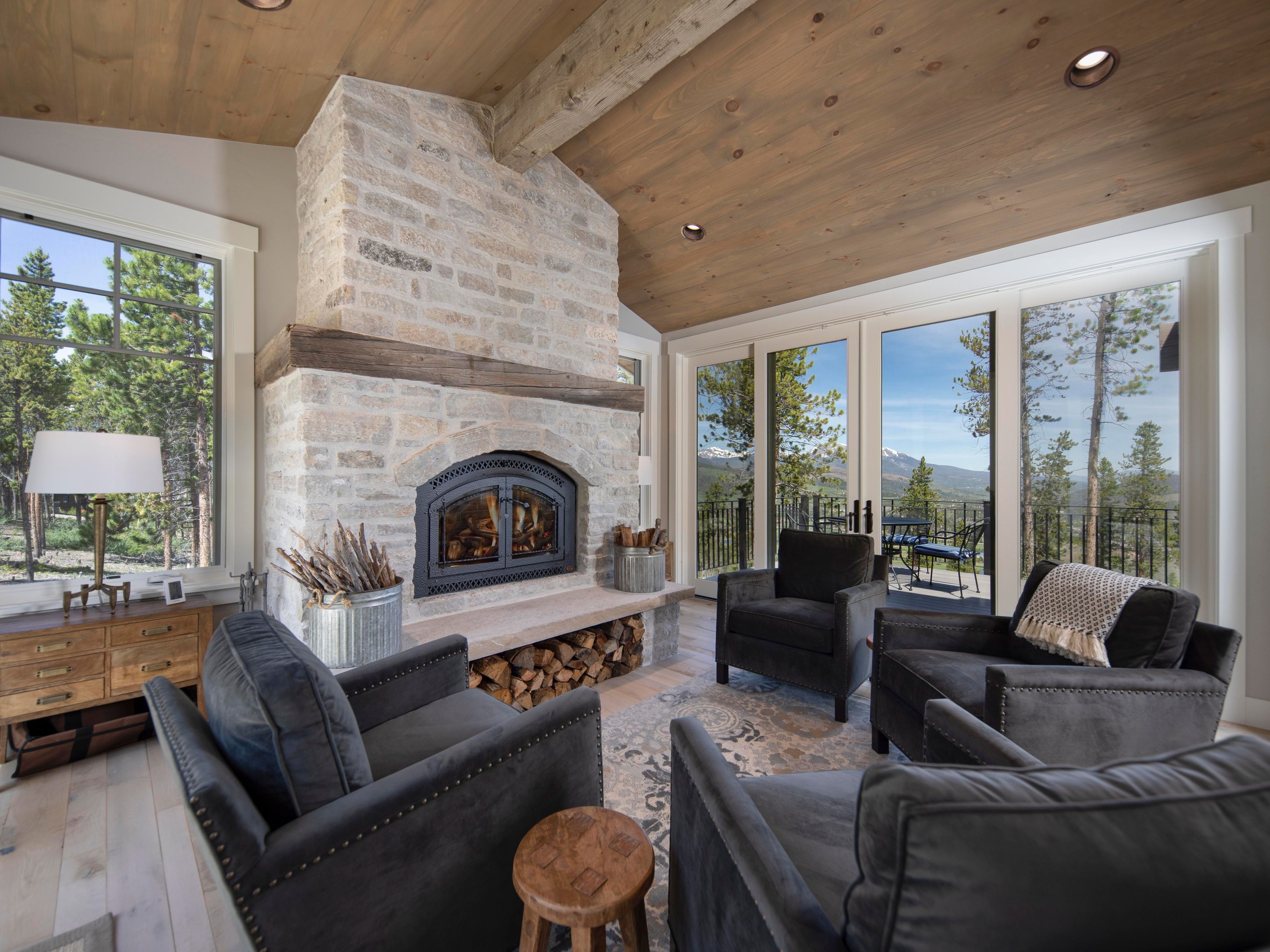 This cozy living room features a prominent stone-clad chimney breast with a built-in fireplace and a rustic wood mantel. The space is furnished with four dark upholstered armchairs arranged around a patterned area rug, creating an intimate seating area. Large glass doors provide expansive views of the surrounding forest and distant mountains, while the vaulted wood-paneled ceiling adds a warm, cabin-like aesthetic to the room.