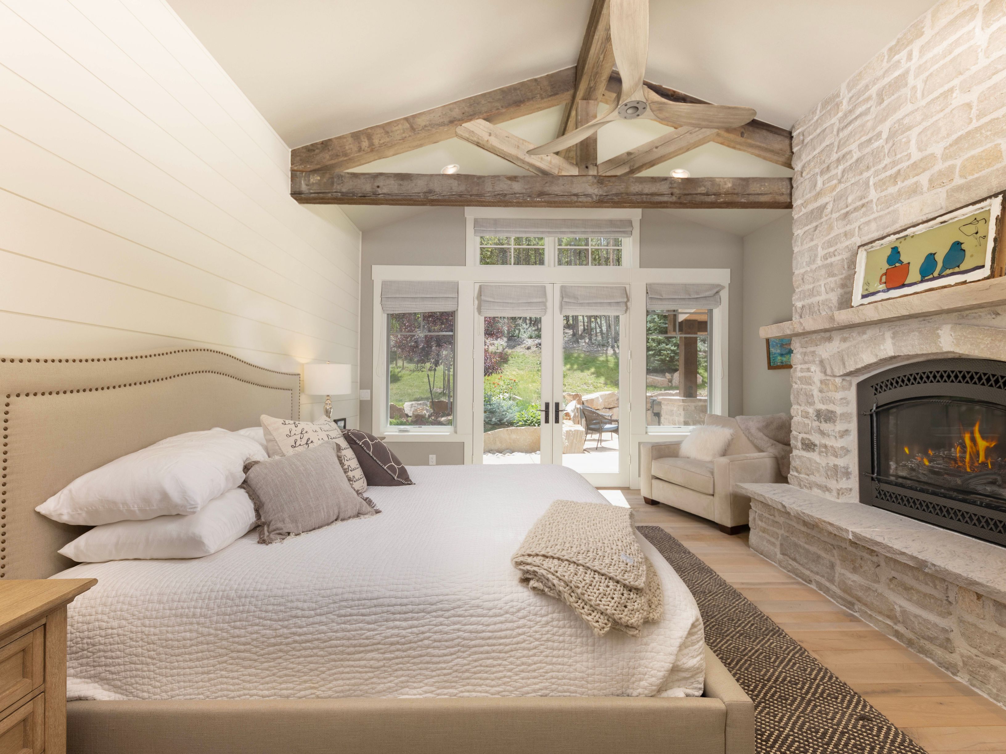 This primary bedroom features a vaulted ceiling with exposed wooden beams and a central ceiling fan, creating a rustic yet refined atmosphere. A prominent stone-clad chimney breast houses a fireplace, while a large upholstered bed sits opposite a set of glass doors that open to a scenic outdoor view. The room is finished with light-toned shiplap walls, a neutral color palette, and a cozy seating area, offering a serene and luxurious retreat.