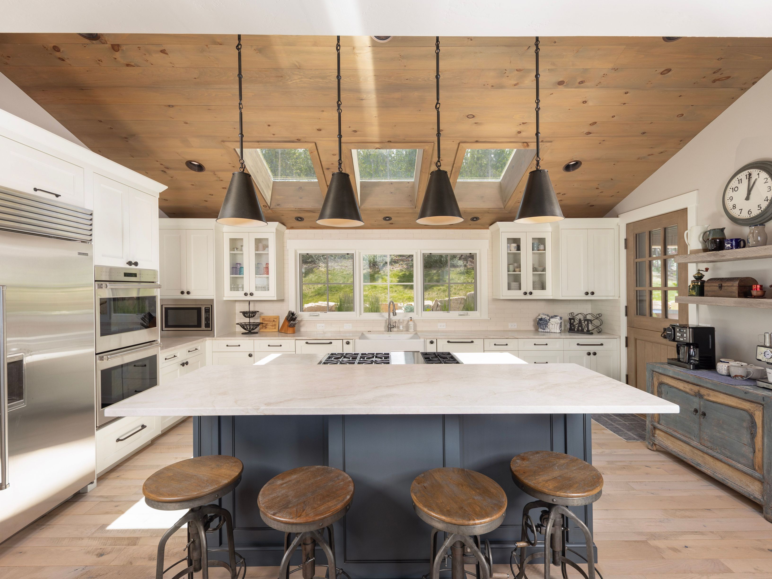 This bright, farmhouse-inspired kitchen features a large central island with a stone countertop and dark cabinetry, complemented by four industrial-style stools. The space is defined by a vaulted wood-paneled ceiling with integrated skylights and four pendant lights hanging over the island. White perimeter cabinetry, stainless steel appliances, and a window-lined wall create an airy, functional, and inviting atmosphere.