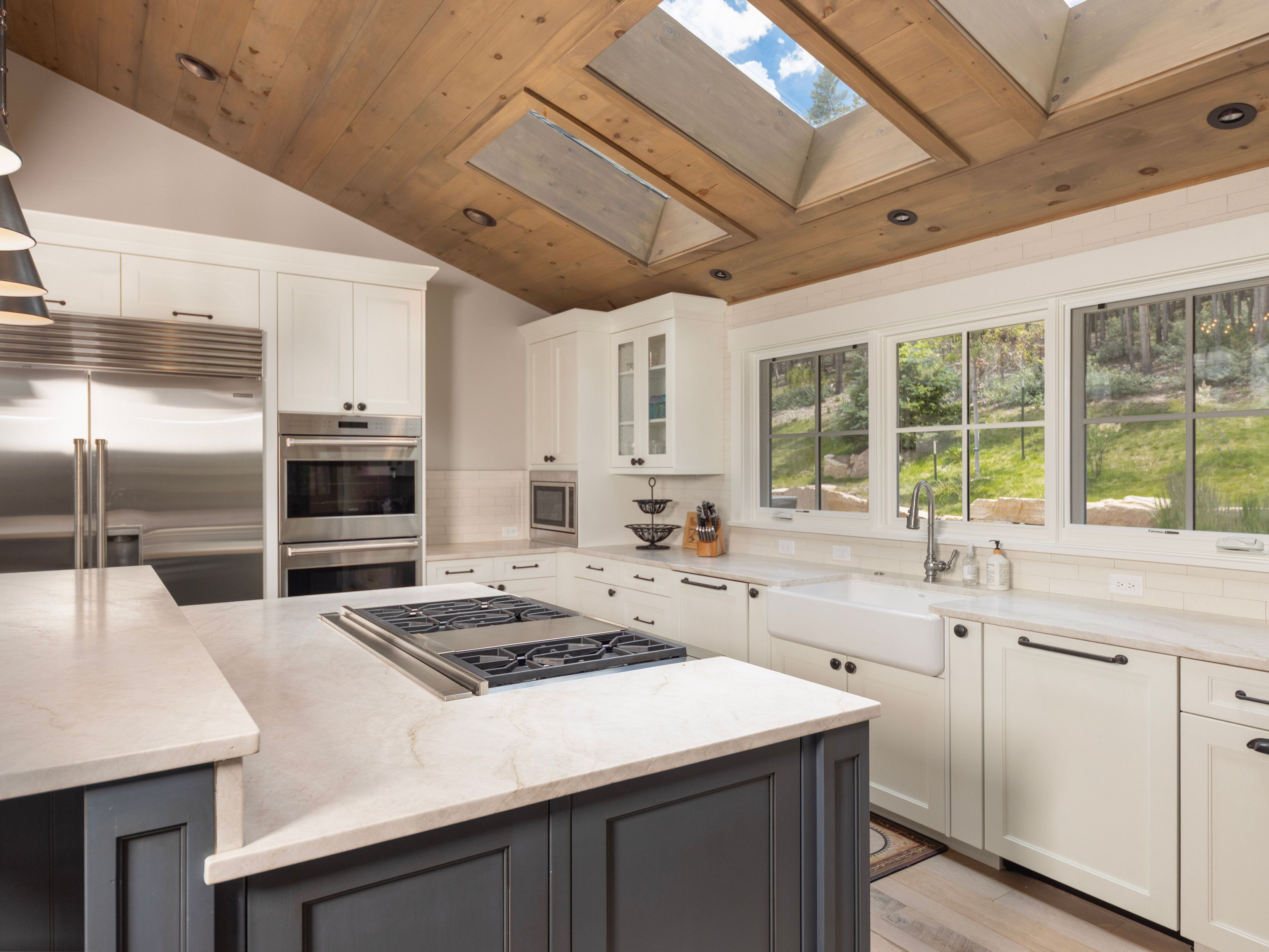 This bright and airy kitchen features a large central island with a gas cooktop and a contrasting dark base, complemented by white perimeter cabinetry and light-toned stone countertops. The space is defined by a vaulted wood-paneled ceiling with integrated skylights that flood the room with natural light, while a farmhouse-style sink sits beneath a row of windows overlooking the outdoors. The overall aesthetic is a blend of rustic warmth and modern functionality, creating an inviting atmosphere for cooking and gathering.