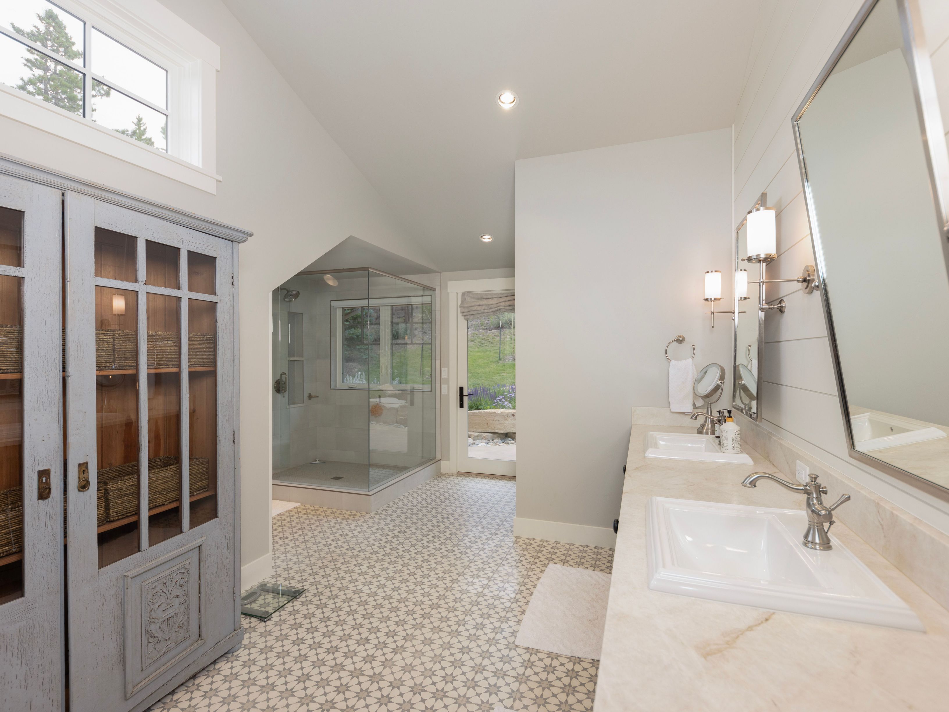 This spacious primary bathroom features a double vanity with a light-toned stone countertop and dual sinks, complemented by a large mirror and wall-mounted lighting. A glass-enclosed walk-in shower is positioned in the background, while a tall, light-blue cabinet with glass-paneled doors provides elegant storage. The room is finished with patterned floor tiles and a neutral color palette, creating a bright and airy atmosphere with a view leading toward an exterior door.