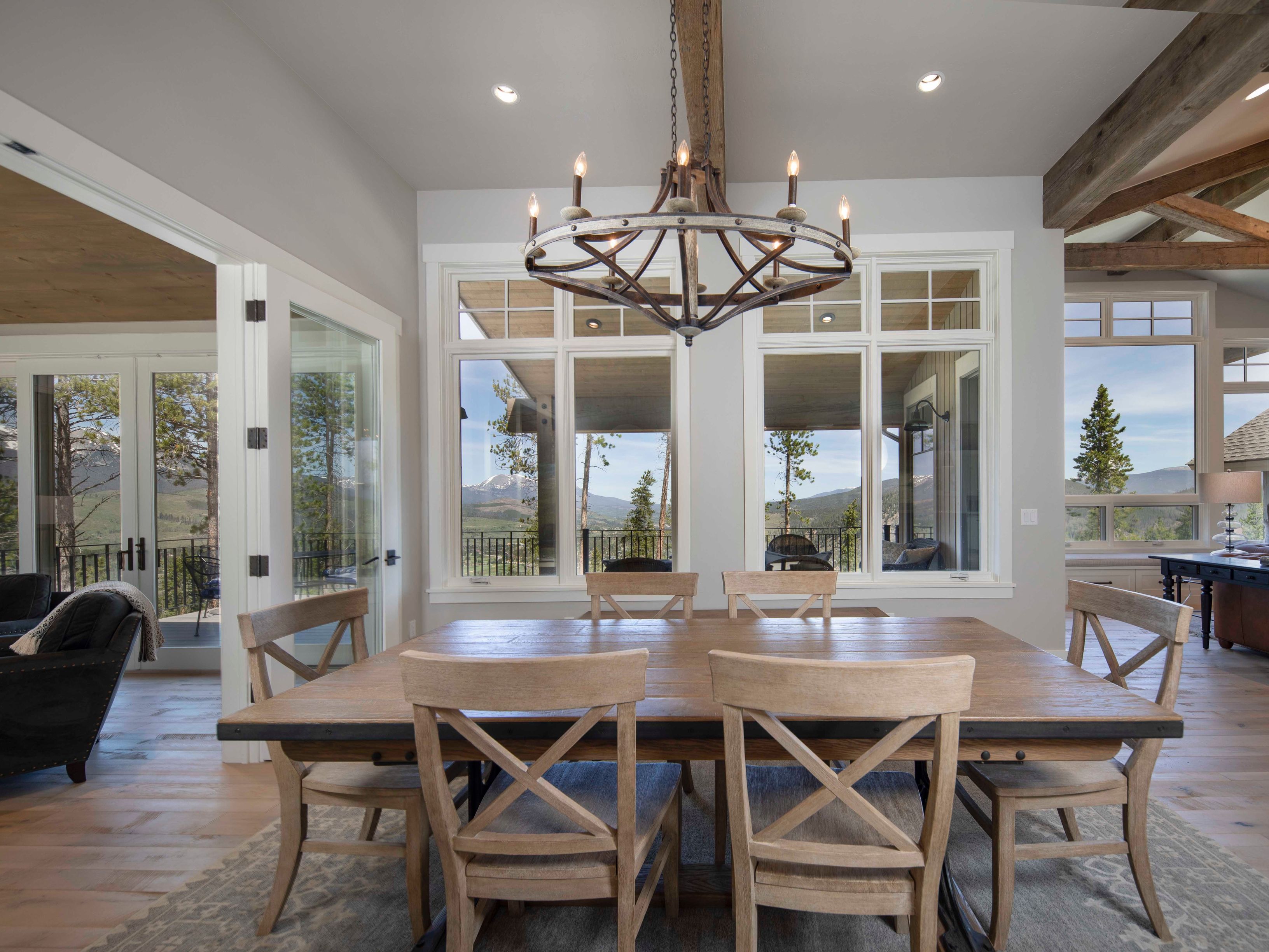 This elegant dining room features a large wooden table surrounded by six light-wood chairs with cross-back detailing, set atop a patterned area rug. A rustic, wagon-wheel style chandelier hangs from a wooden beam, serving as the focal point above the table. Large windows provide an expansive view of the mountainous landscape outside, while an open doorway to the left reveals a glimpse of a comfortable living area.