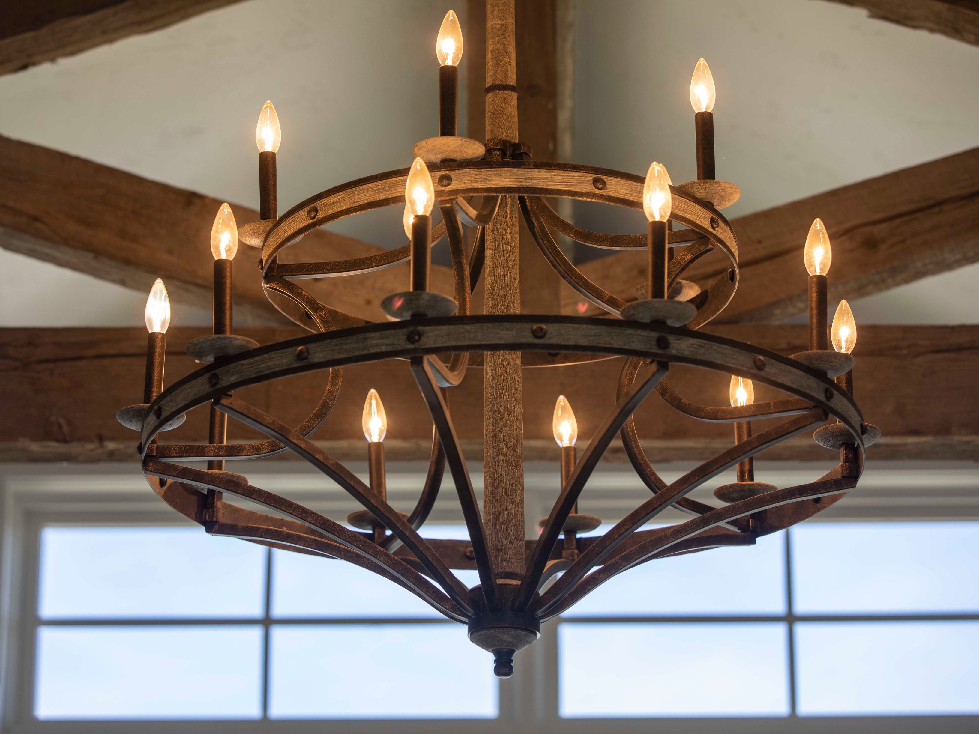This image features a large, rustic-style chandelier with a two-tiered circular frame and a central wooden post. The fixture is equipped with numerous candle-style bulbs that provide warm, ambient lighting, set against a backdrop of exposed wooden ceiling beams and a window with a grid pattern. The perspective is a low-angle, centered shot that emphasizes the intricate metalwork and the warm, inviting glow of the light fixture.