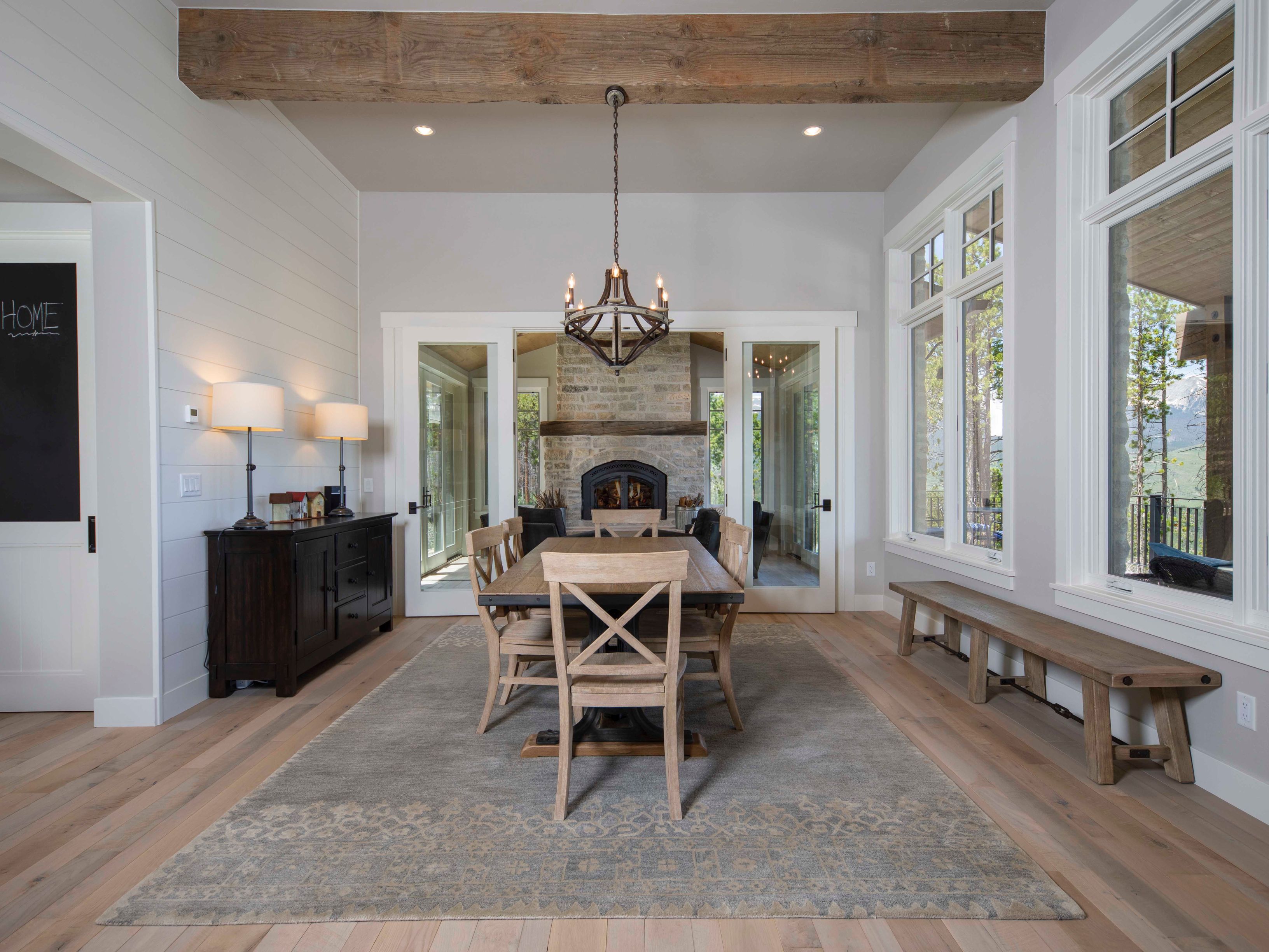 This elegant dining room features a central wooden table surrounded by cross-back chairs, set atop a large, muted-pattern area rug. A dark wood sideboard with matching lamps sits against a white shiplap wall, while a rustic wooden beam spans the ceiling above a chandelier. Through the glass-paned doors, a stone-clad chimney breast is visible in the adjacent room, and large windows on the right provide ample natural light and a view of the outdoors.