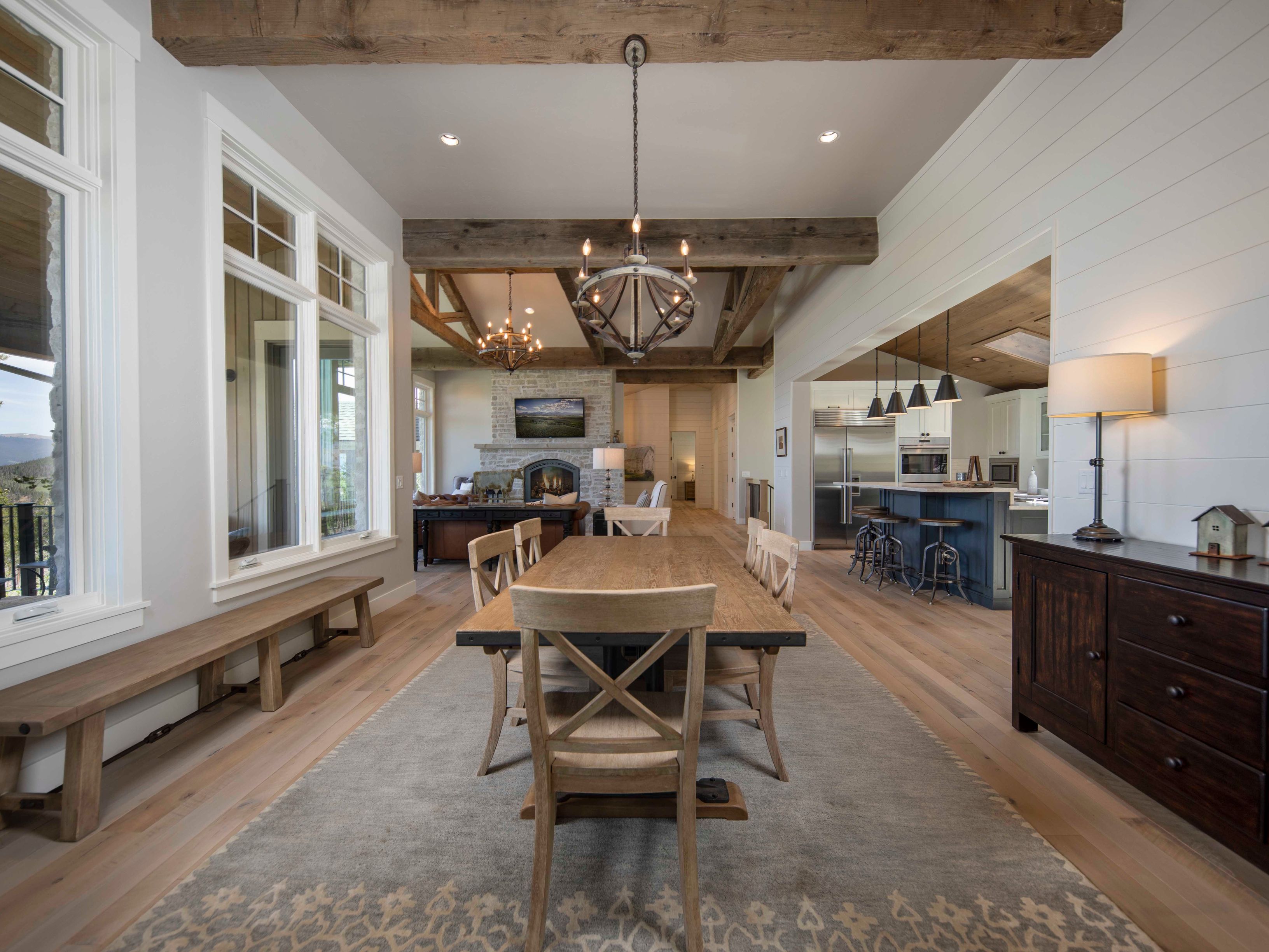 This open-concept dining area features a rustic wooden table surrounded by cross-back chairs, set atop a large patterned area rug. The space is defined by exposed wooden ceiling beams and a neutral color palette, with a view extending into a kitchen with dark cabinetry and a living area anchored by a stone-clad chimney breast. Large windows along the left wall provide ample natural light, creating an inviting and spacious atmosphere suitable for entertaining.