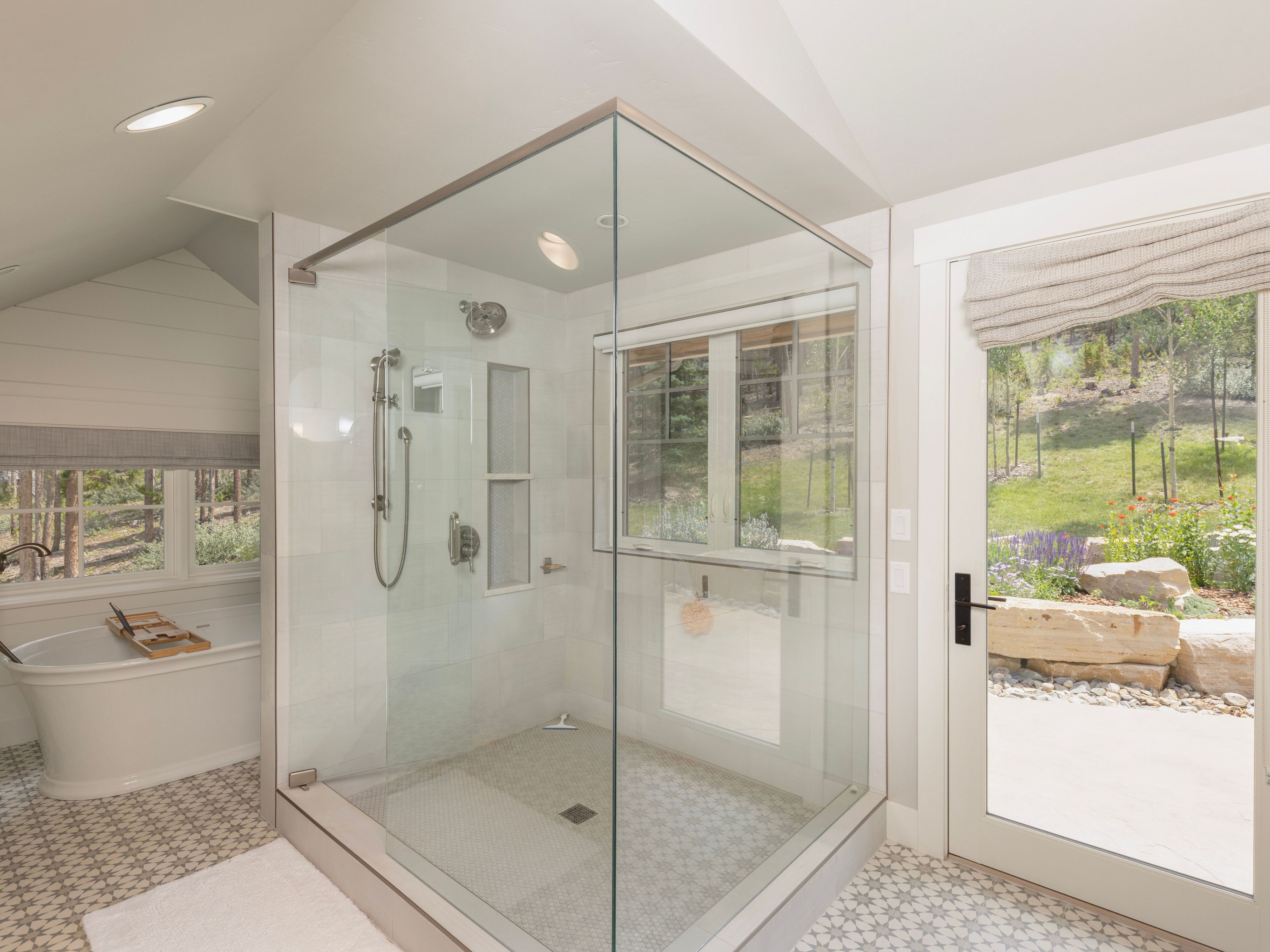 This bright primary bathroom features a large glass-enclosed walk-in shower with patterned tile flooring and a built-in niche. To the left, a freestanding soaking tub sits beneath a window, while a glass door on the right provides direct access to the outdoor garden area. The space is characterized by a clean, neutral aesthetic with light-toned walls and natural light streaming in from multiple windows.