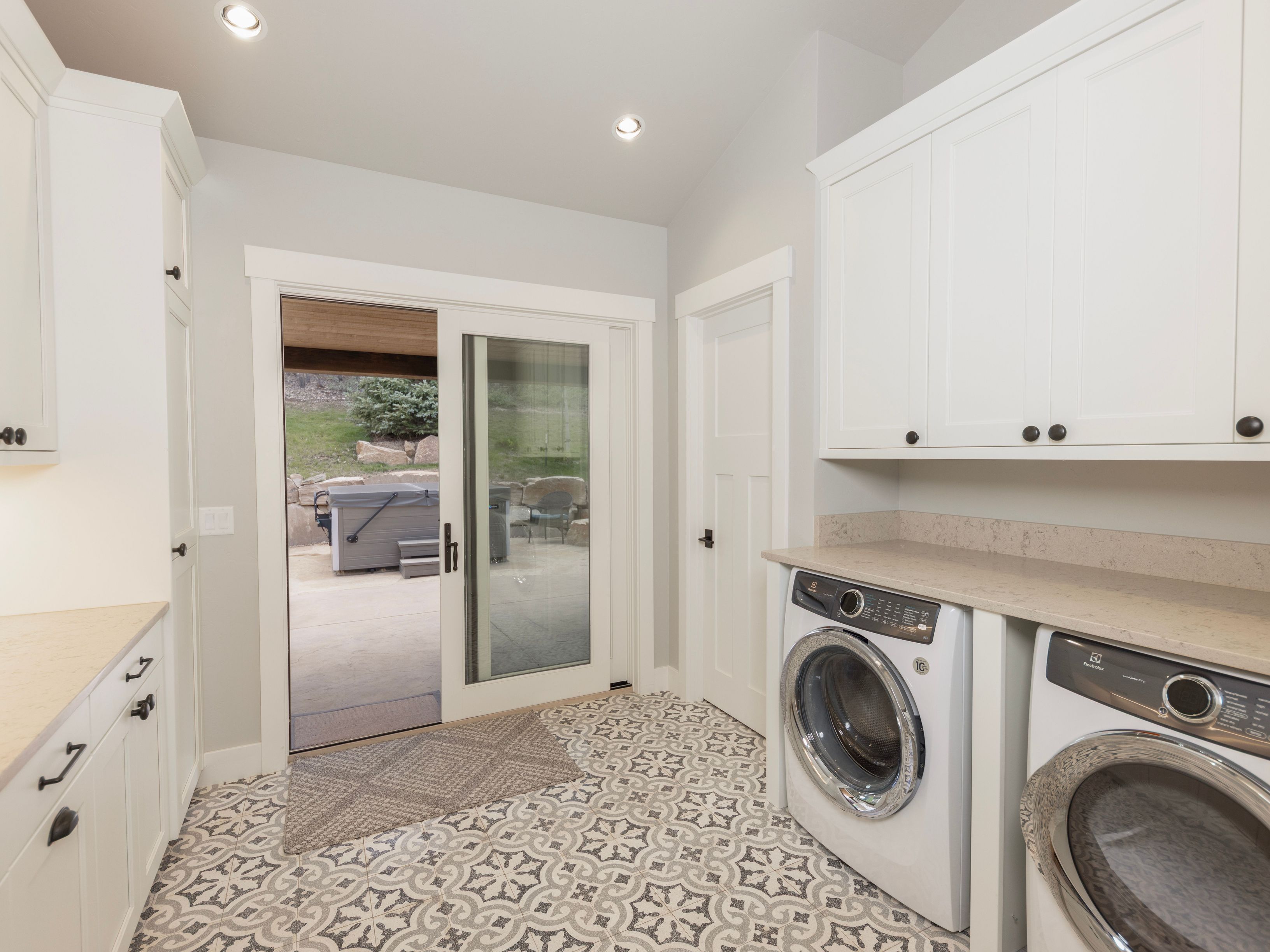 This bright and functional laundry room features white cabinetry, light-toned countertops, and a striking patterned tile floor. A sliding glass door provides a direct view and access to an outdoor patio area with a spa. The space is well-lit with recessed lighting and includes a dedicated laundry area with a washer and dryer.