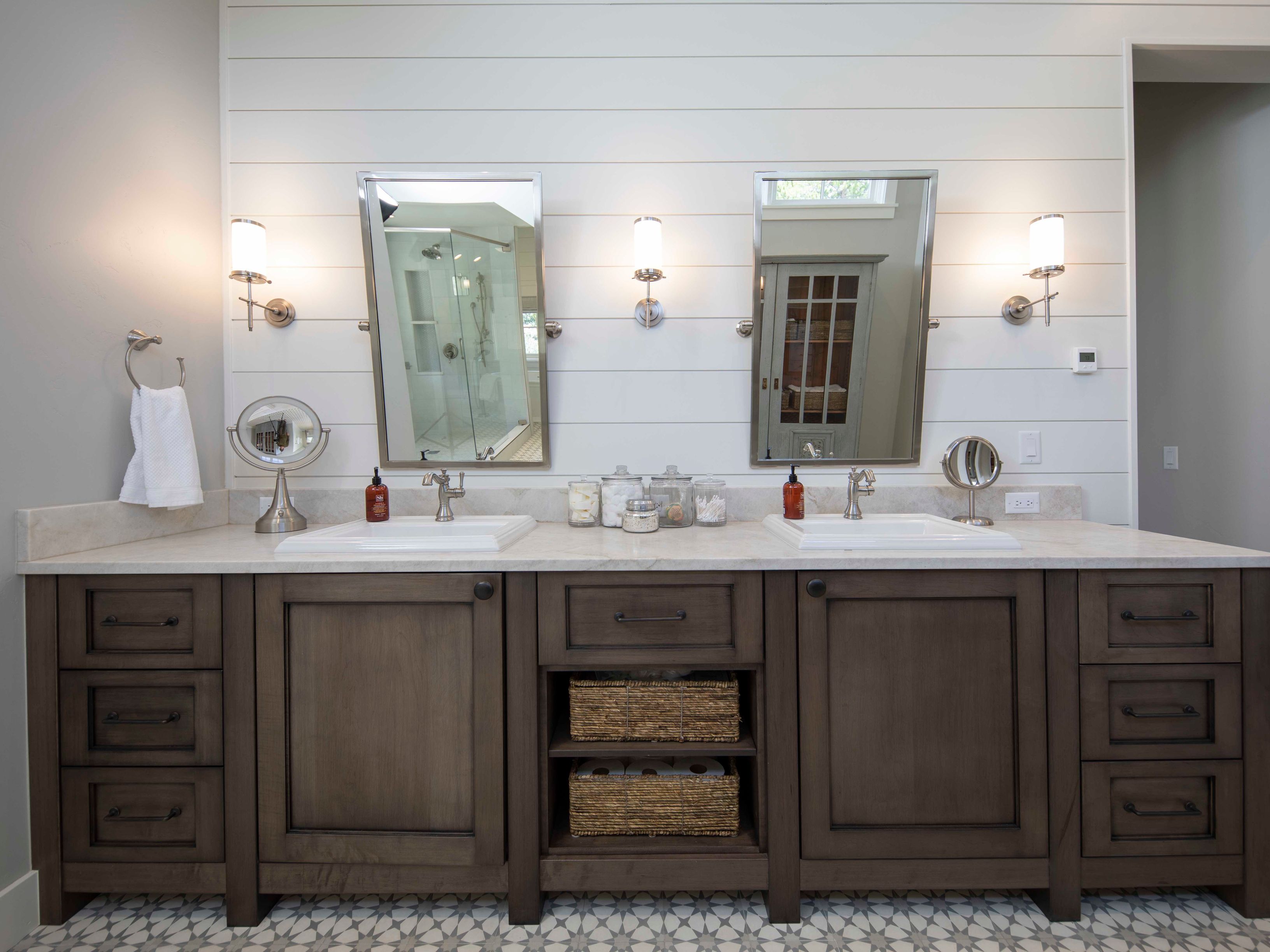 This primary bathroom features a double vanity with dark wood cabinetry, a light-colored stone countertop, and dual rectangular sinks. The wall behind the vanity is finished with horizontal white shiplap, accented by two rectangular mirrors and four wall-mounted light fixtures. Below, the floor is covered in a decorative patterned tile, and the center of the vanity includes open shelving with woven baskets for storage.