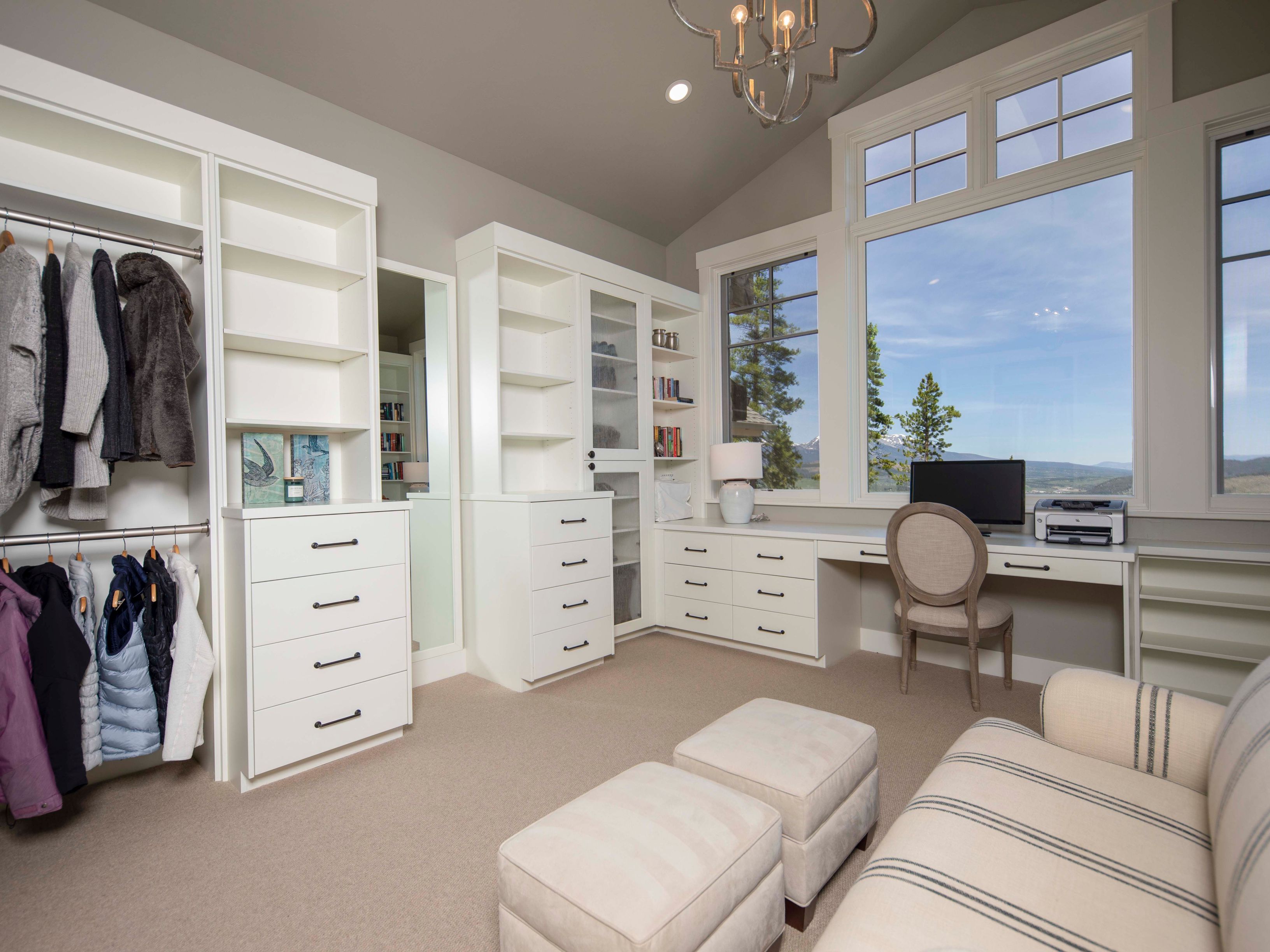 This versatile room functions as both a home office and a walk-in closet, featuring extensive white built-in cabinetry, shelving, and a dedicated desk area. Large windows provide abundant natural light and expansive mountain views, while a neutral color palette and soft carpeting create a serene atmosphere. The space is furnished with a comfortable chair, two ottomans, and a striped sofa, making it a functional and inviting multi-purpose area.