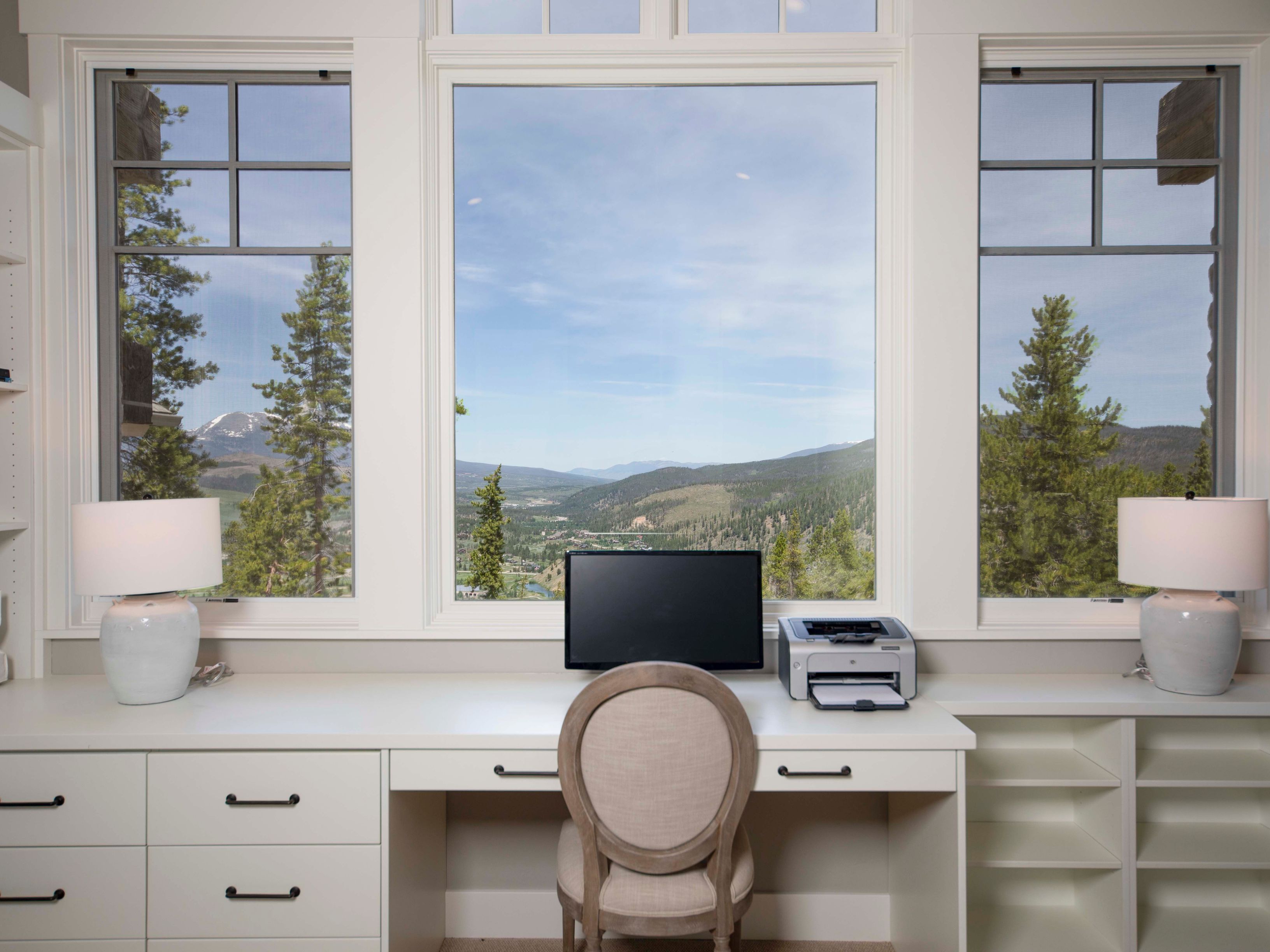 This bright home office features a built-in white desk setup positioned directly in front of a large, multi-pane window that frames a scenic mountain view. The workspace includes a central computer monitor, a printer, and matching table lamps on either side, complemented by a neutral-toned upholstered chair. The clean, minimalist design is enhanced by ample drawer storage and open shelving, creating a serene and productive environment.