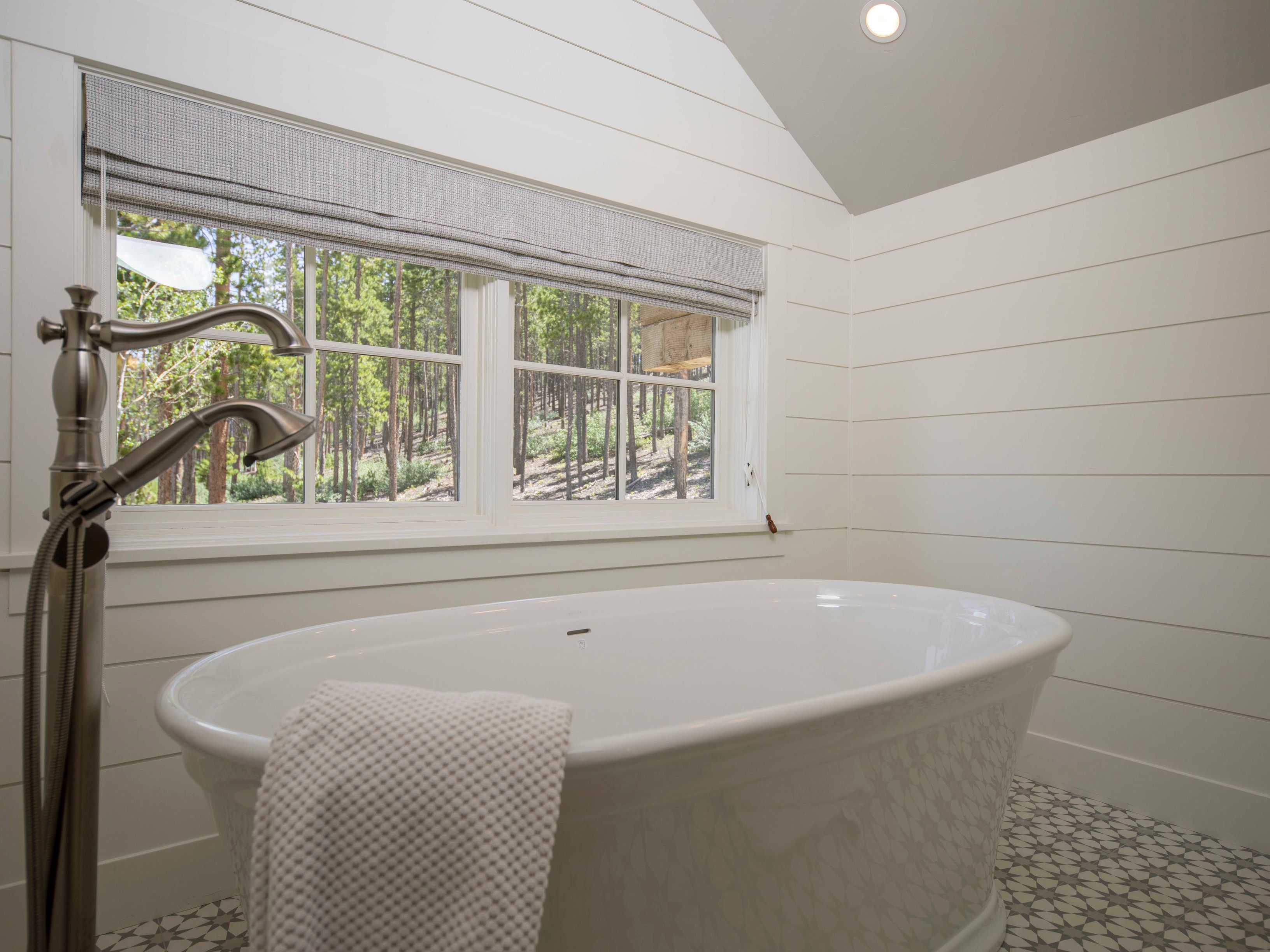 This serene bathroom features a freestanding soaking tub positioned beneath a window that offers a peaceful view of a wooded landscape. The walls are finished with white horizontal shiplap, complemented by patterned floor tiles and a sleek, metallic floor-mounted tub filler. The vaulted ceiling and natural light create an airy, spa-like atmosphere.
