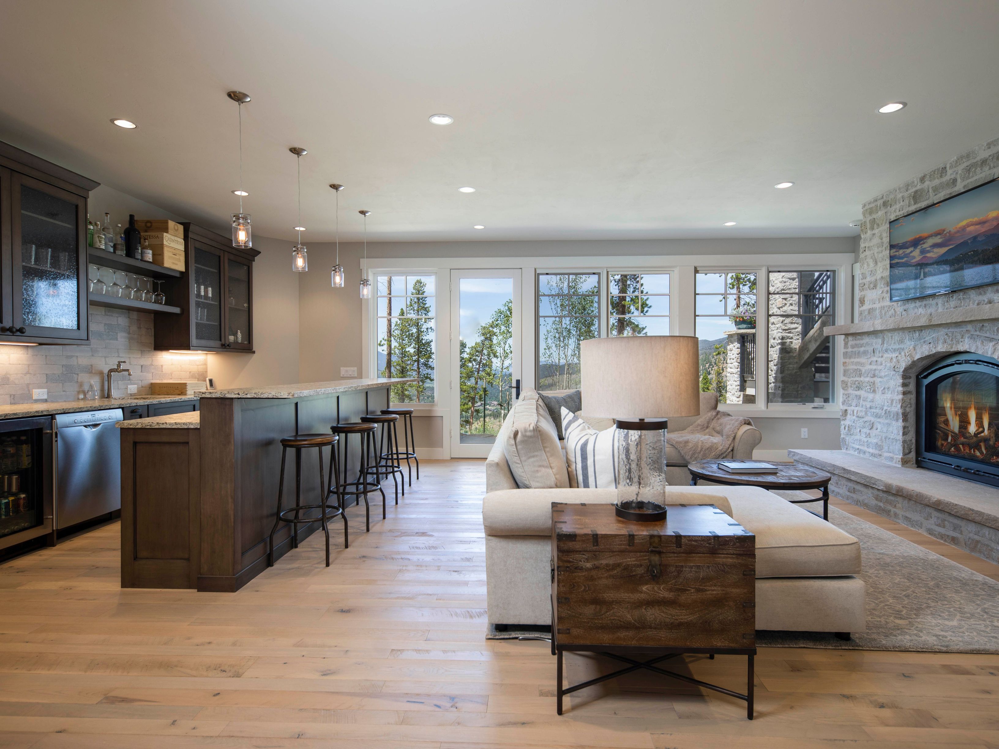 This open-concept living space seamlessly blends a kitchen area with a cozy lounge, featuring light wood flooring and a neutral color palette. The kitchen is equipped with dark cabinetry, a stone-topped island with bar seating, and pendant lighting, while the adjacent living area centers around a stone-clad chimney breast with an integrated fireplace and television. Large windows and a glass door provide abundant natural light and views of the surrounding trees, creating a warm and inviting atmosphere.