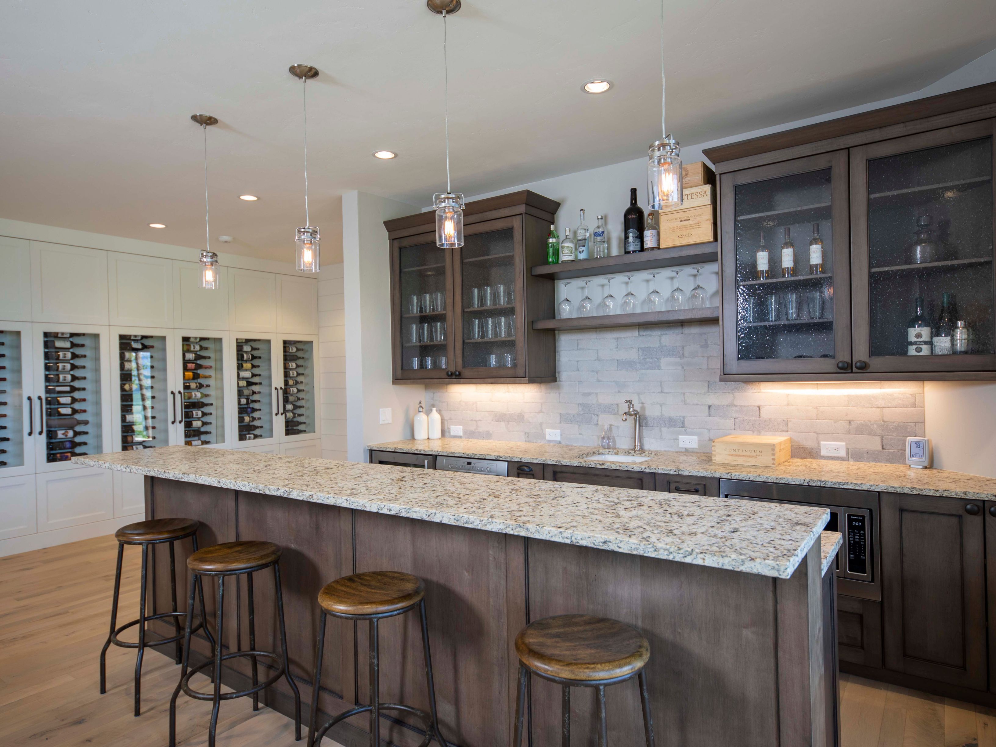 This sophisticated home bar area features a long island with a stone countertop and four rustic stools, perfect for entertaining. The space is anchored by dark wood cabinetry, a light-toned brick backsplash, and a dedicated wine storage wall with glass-fronted cabinets. The perspective is a wide, eye-level shot that captures the warm, inviting atmosphere of this high-end wet bar.