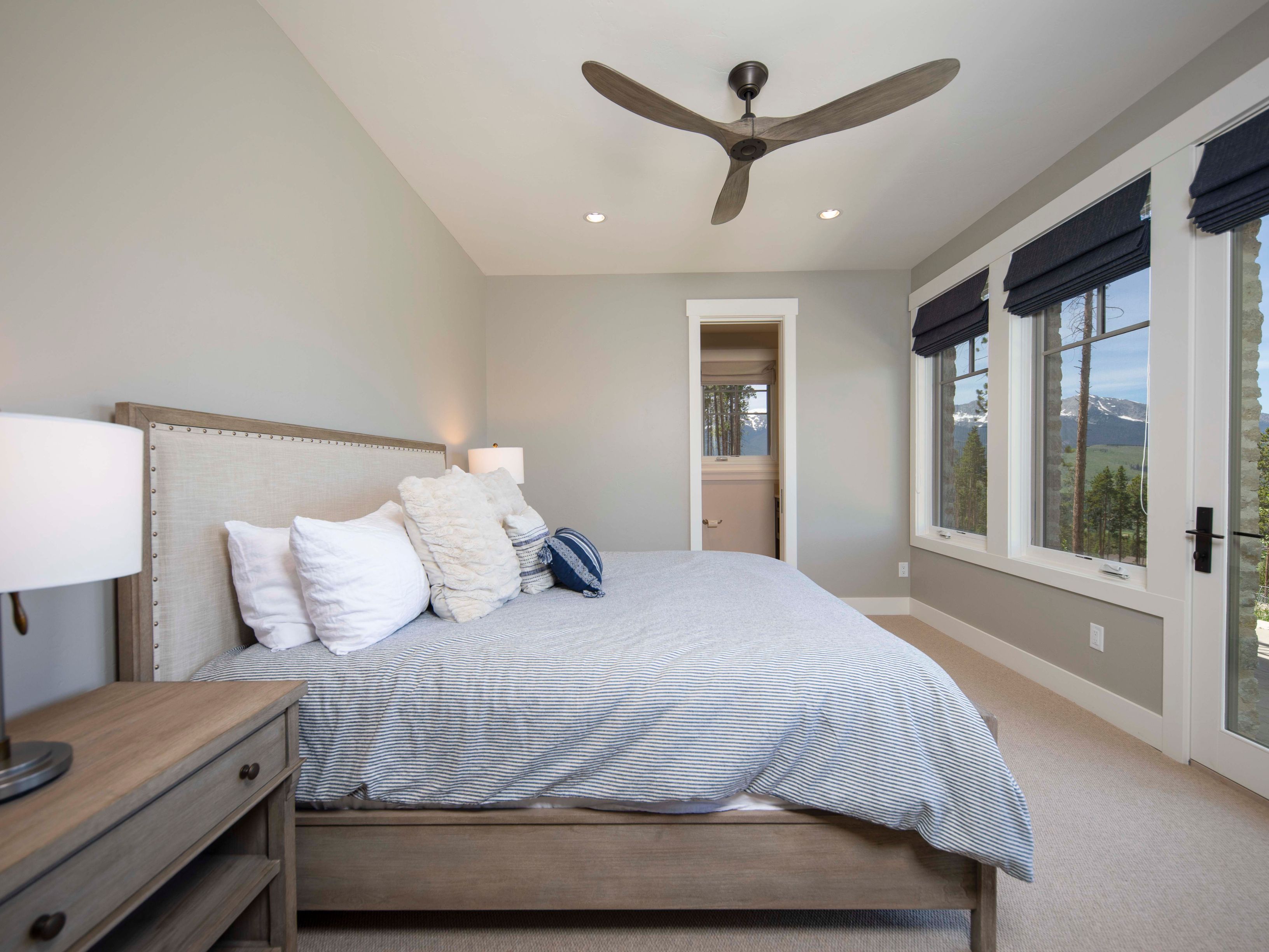 This serene primary bedroom features a neutral color palette with light gray walls and a light wood bed frame topped with a striped duvet and plush pillows. Large windows with dark blue roman shades provide ample natural light and scenic mountain views, while a ceiling fan hangs centrally above. A bedside table with a lamp sits to the left, and a doorway leads to an adjacent room, creating an open and inviting atmosphere.