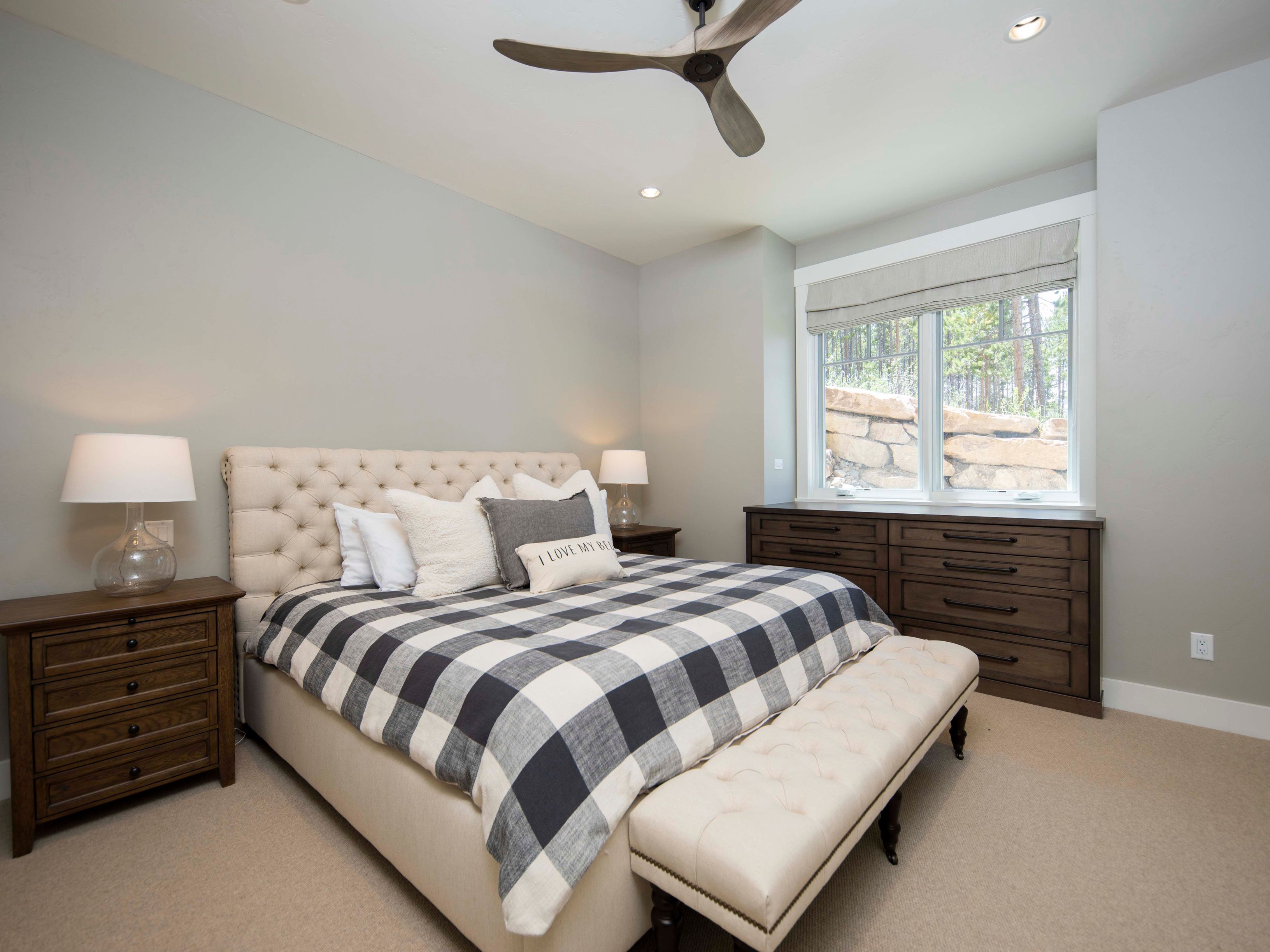 This primary bedroom features a neutral color palette with a tufted upholstered headboard and a classic black-and-white buffalo check duvet. A matching tufted bench sits at the foot of the bed, while dark wood nightstands and a coordinating dresser provide ample storage. The room is illuminated by a modern ceiling fan and soft bedside lamps, with a window offering a view of the natural landscape outside.