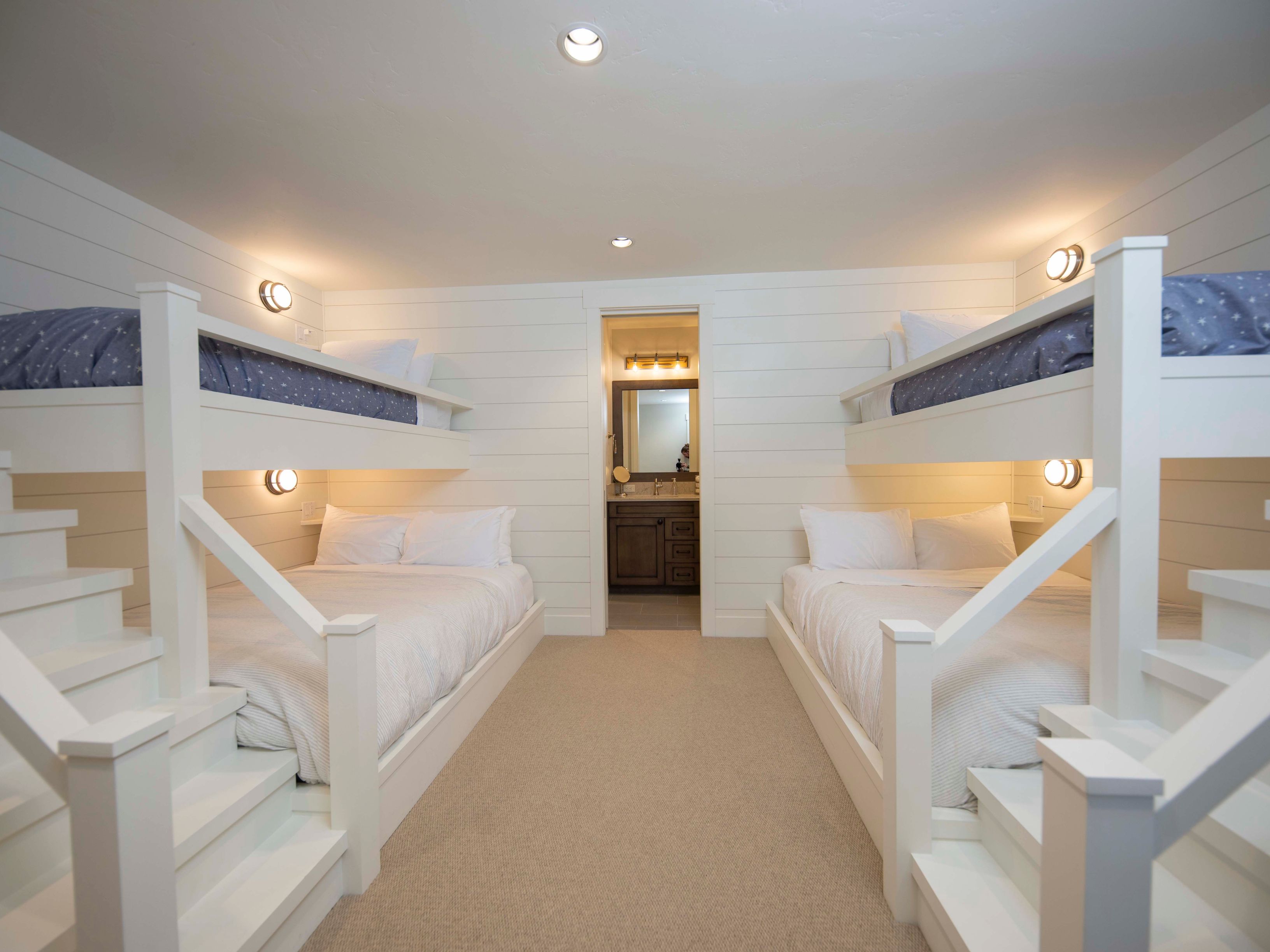This bedroom features a symmetrical layout with two sets of built-in bunk beds, each equipped with its own staircase for easy access to the upper bunks. The room is finished with white shiplap walls, neutral carpeting, and warm wall-mounted lighting, creating a cozy and functional sleeping space. A central doorway leads directly into a well-lit bathroom, enhancing the convenience of this guest suite.