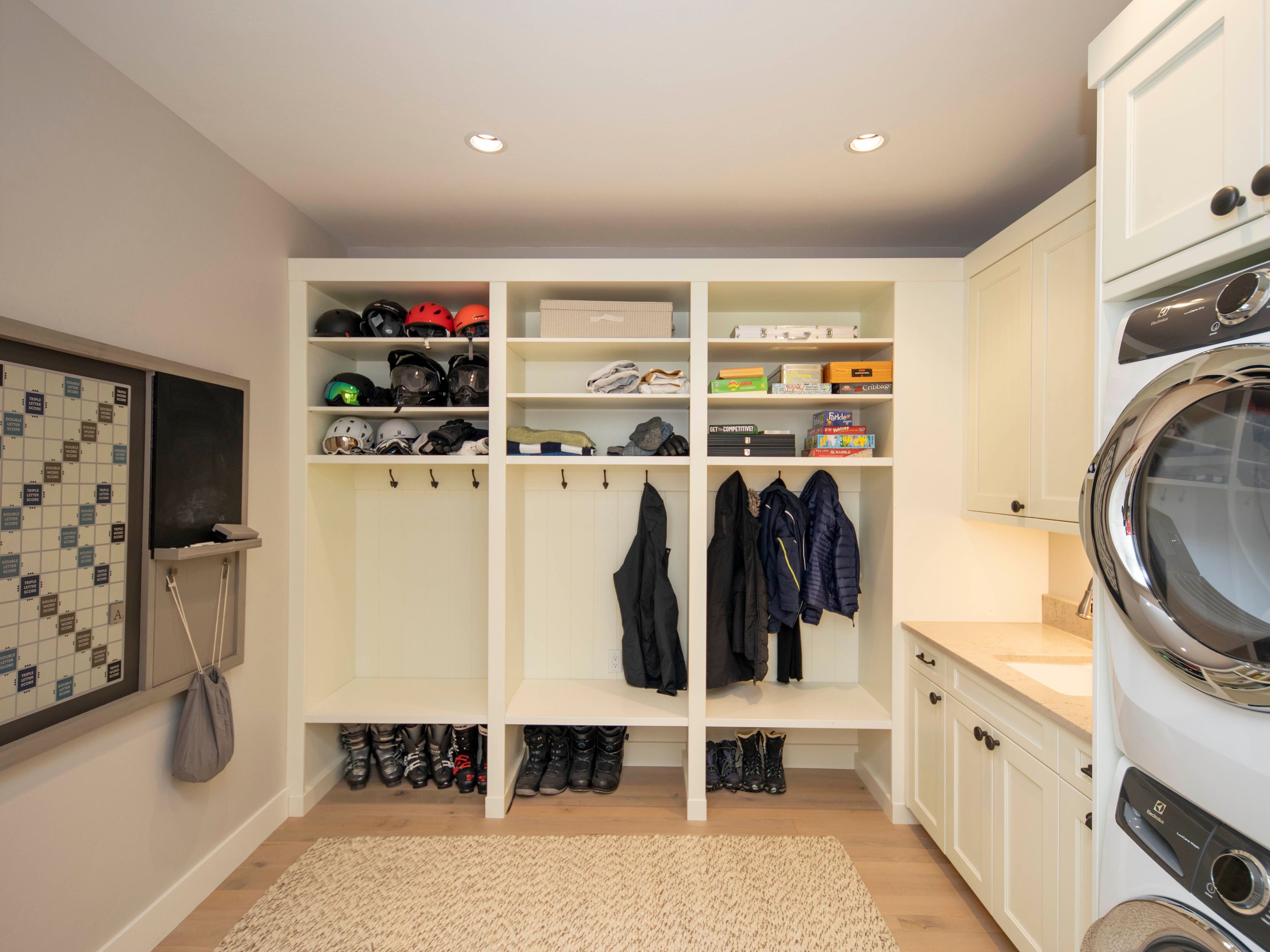 This organized laundry and mudroom area features a large built-in storage unit with multiple cubbies for helmets, coats, and footwear. To the right, a stacked washer and dryer set is integrated into cabinetry with a light-toned countertop and sink area. The space is finished with light wood flooring and a neutral rug, creating a functional and tidy transition zone.