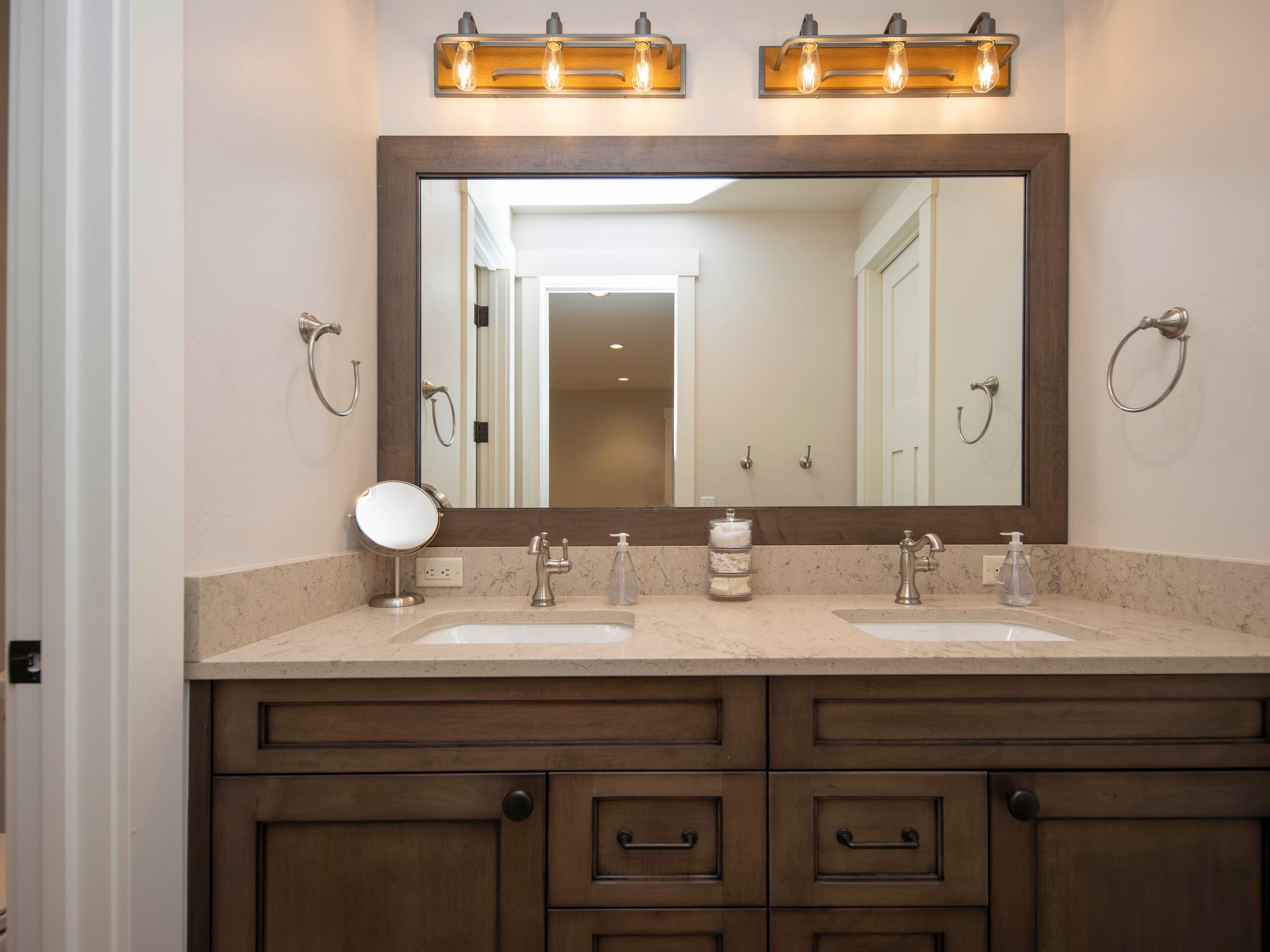 This double-vanity bathroom features a dark wood cabinet base topped with a light-toned stone countertop and dual undermount sinks. A large, rectangular mirror with a matching dark wood frame is centered above the vanity, flanked by two industrial-style light fixtures with exposed bulbs. The space is finished with brushed nickel hardware, including towel rings and faucets, creating a cohesive and functional aesthetic.