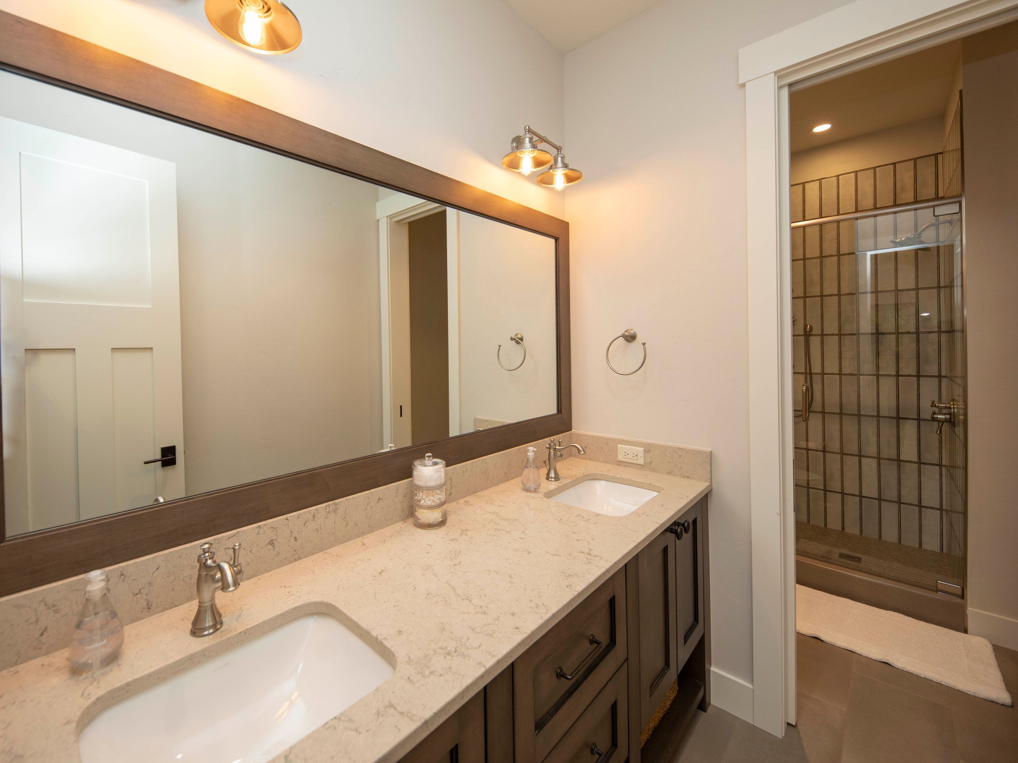 This primary bathroom features a double vanity with a light-toned stone countertop and dark wood cabinetry. A large, wood-framed mirror spans the length of the vanity, complemented by warm-toned wall lighting. To the right, a glass-enclosed shower with vertical tile work is visible, creating a clean and functional space.