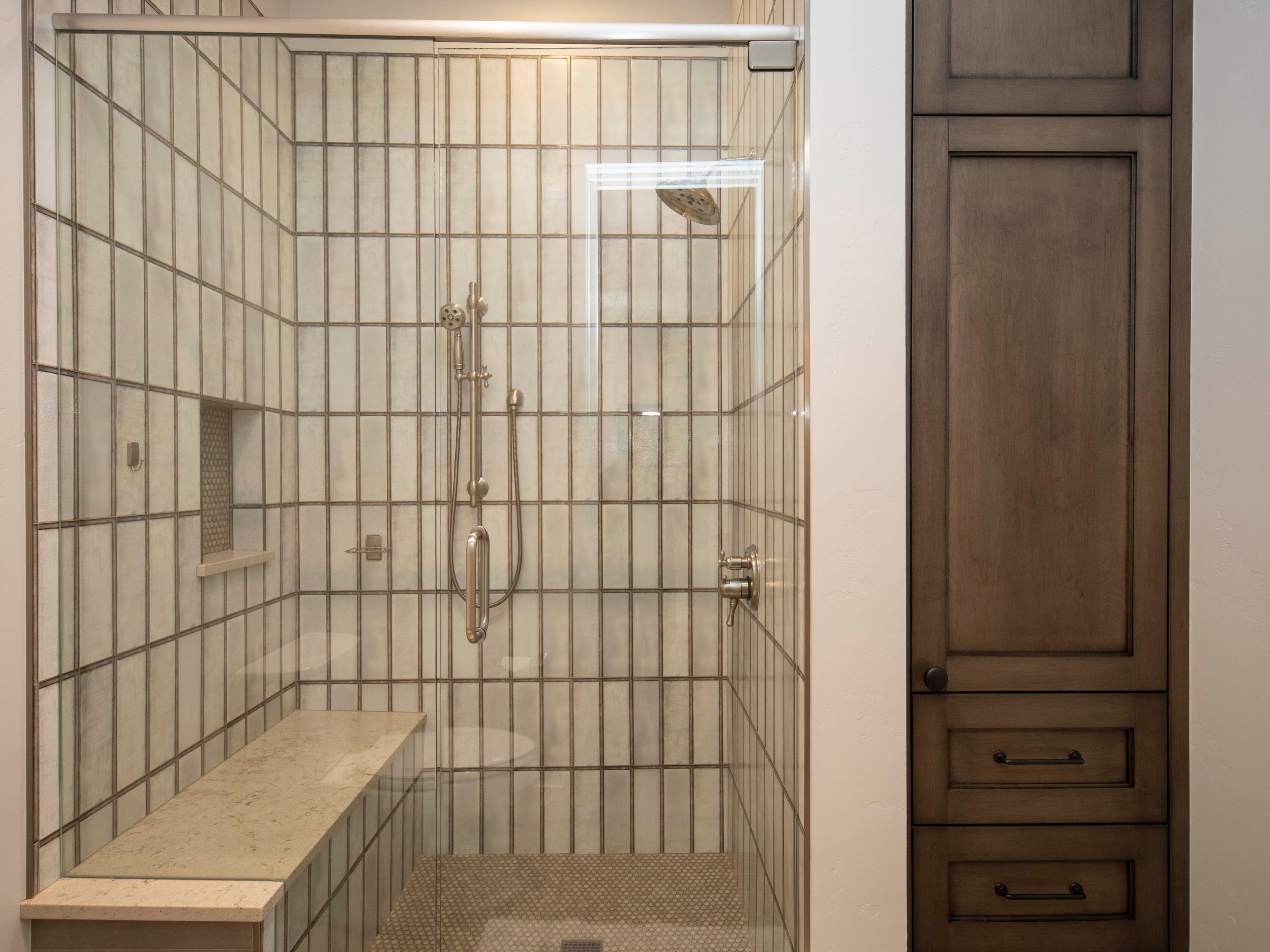This primary bathroom features a walk-in shower enclosed by a glass door, showcasing vertical rectangular wall tiles and a built-in stone bench. To the right, a tall, dark-toned wood cabinet provides integrated storage with drawers and a cabinet door. The shower includes a handheld sprayer and a rainfall showerhead, creating a clean and functional spa-like aesthetic.