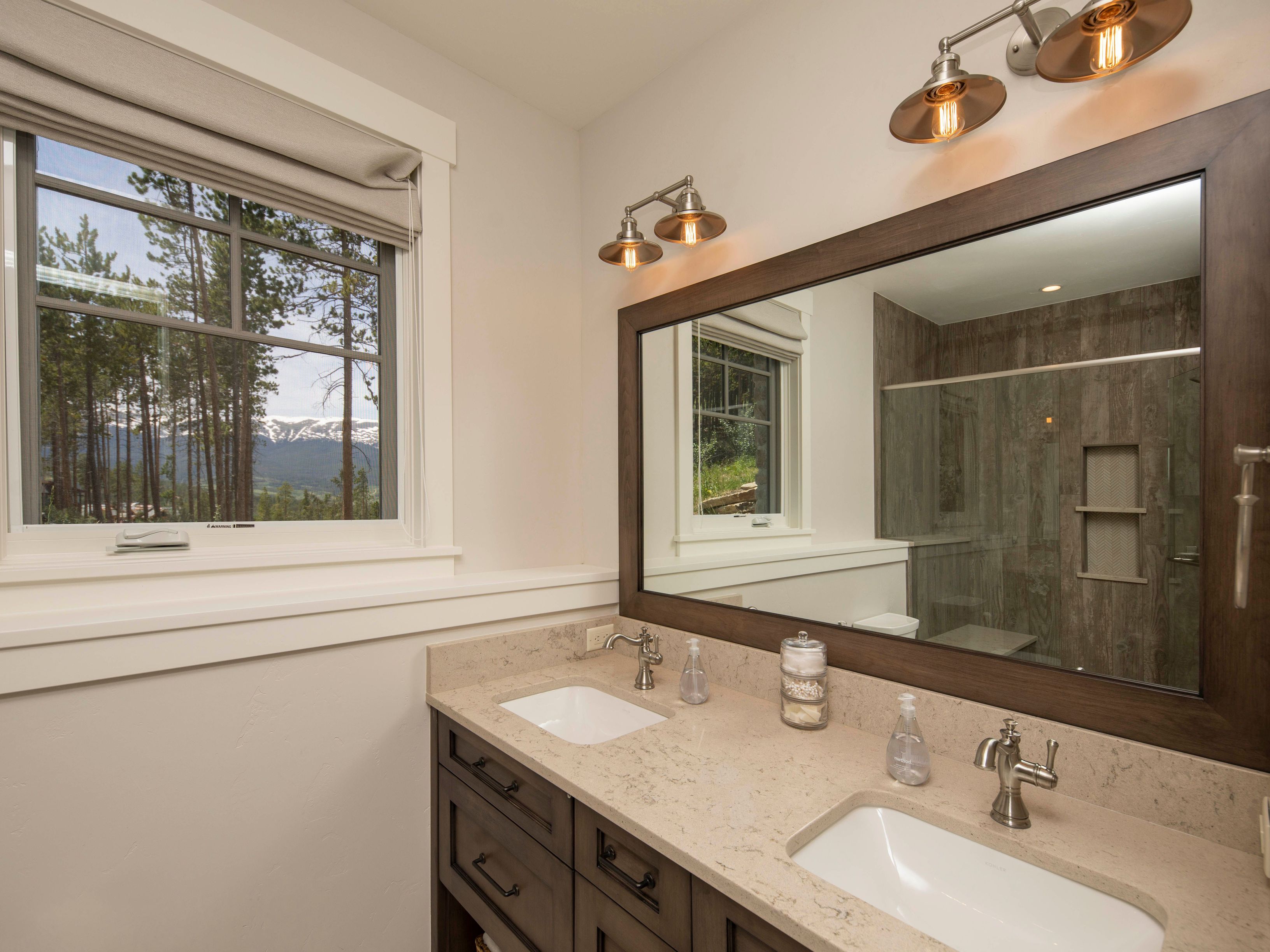 This primary bathroom features a double vanity with a light-toned stone countertop and dark wood cabinetry, complemented by a large mirror framed in matching dark wood. Two sets of industrial-style wall-mounted light fixtures provide warm illumination, while a window to the left offers a scenic view of pine trees and distant snow-capped mountains. The space includes a glass-enclosed shower area with wood-look tile walls, creating a cohesive and rustic-modern aesthetic.