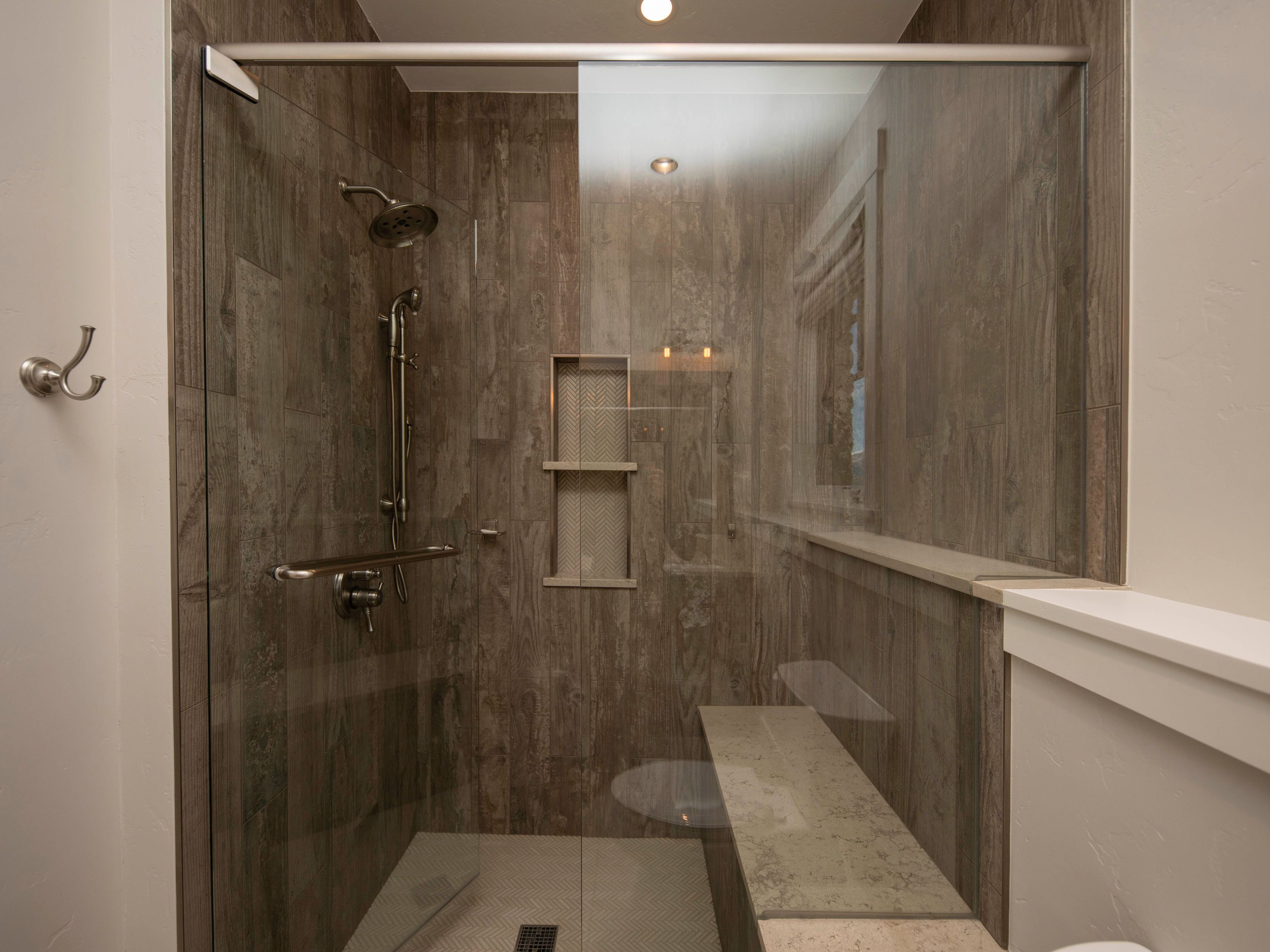 This modern walk-in shower features large-format, wood-look wall tiles and a built-in stone bench for added comfort. A glass enclosure separates the shower area, which includes a dual-head shower system and a recessed niche with patterned tile accents. The space is illuminated by recessed lighting, creating a clean and spa-like atmosphere.