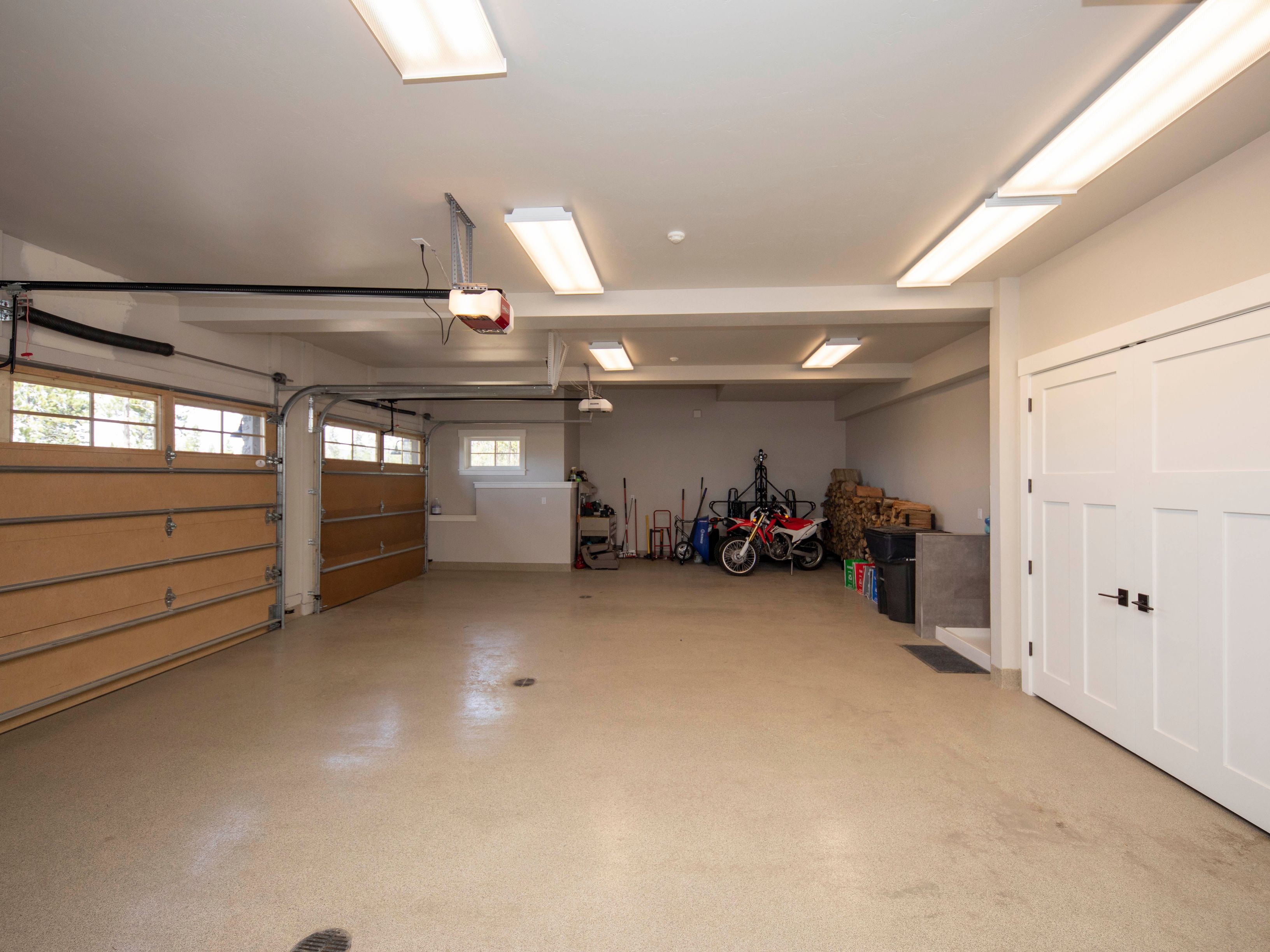 This spacious, well-lit garage features a clean, neutral-toned epoxy floor and multiple overhead fluorescent light fixtures. The area includes two large garage doors with windows, a motorcycle parked in the back, and a stack of firewood, providing a functional and organized storage space. A set of white double doors on the right adds a clean, finished look to the interior.